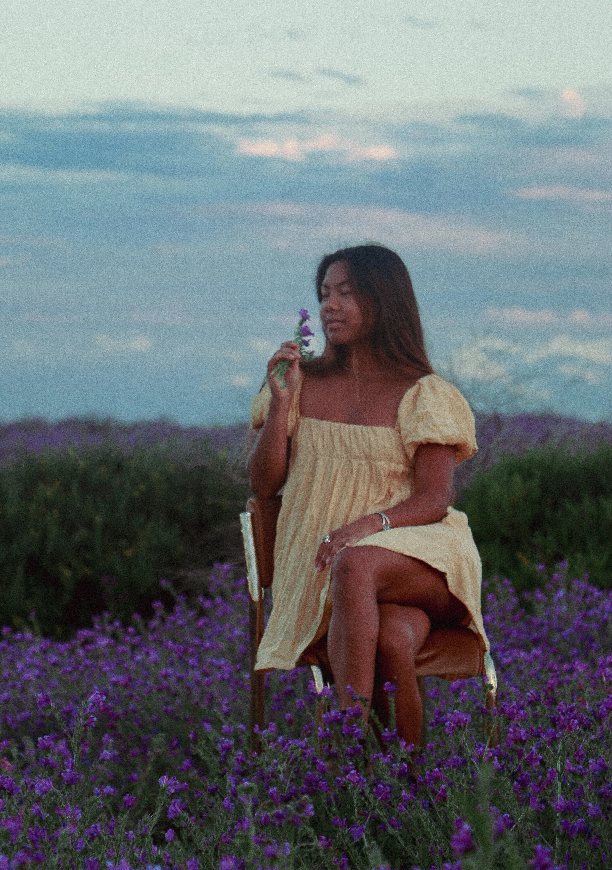 A woman in a yellow dress sitting on a chair in a field of purple flowers, holding a purple flower near her face with her eyes closed at dusk.