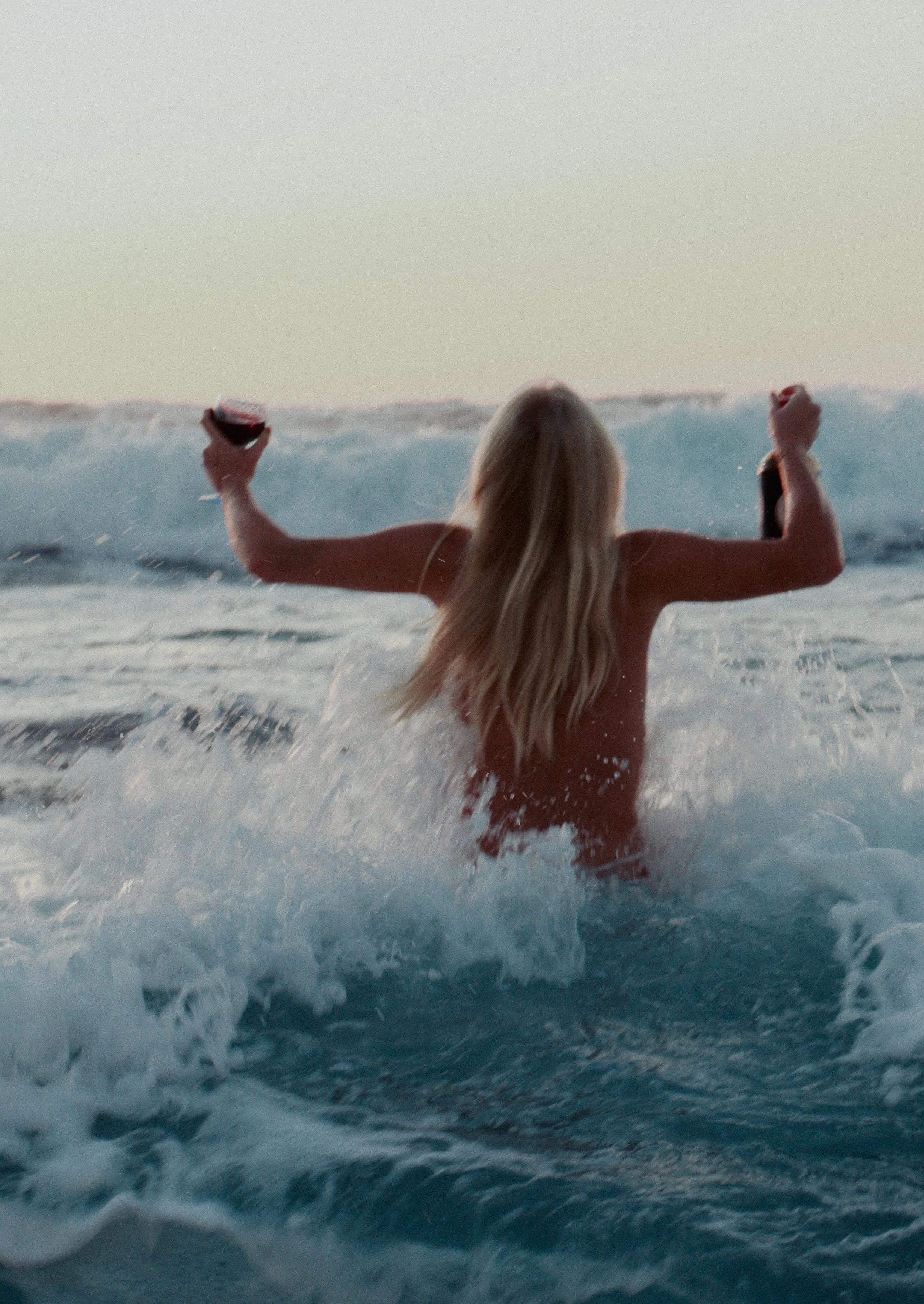 A woman with long blonde hair in the ocean holding a glass of red wine and a small bottle, facing away from the camera, with waves in the background during sunset or sunrise.