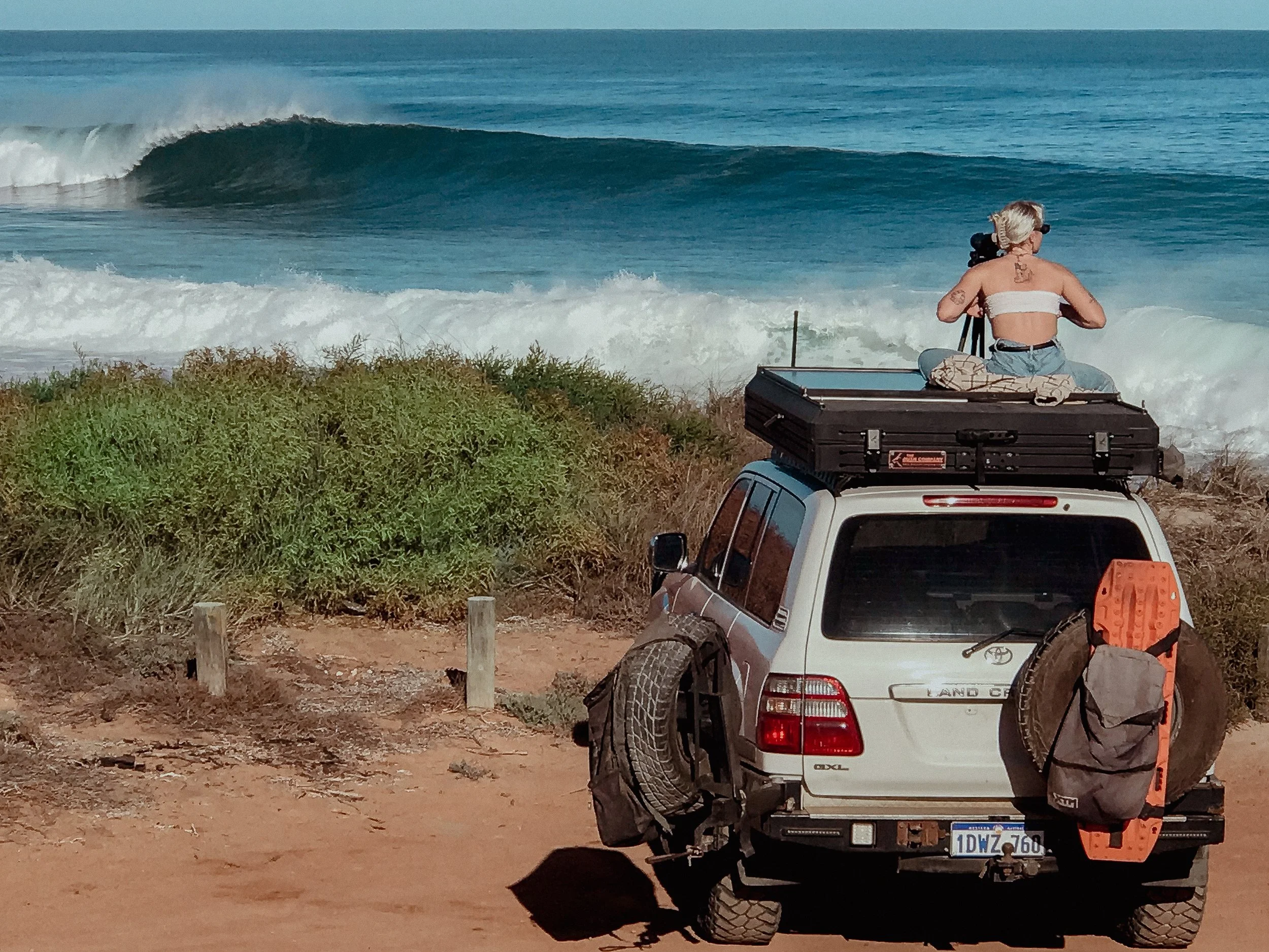 A person sits on top of a white Toyota Land Cruiser parked near the beach, looking through binoculars at the ocean waves.