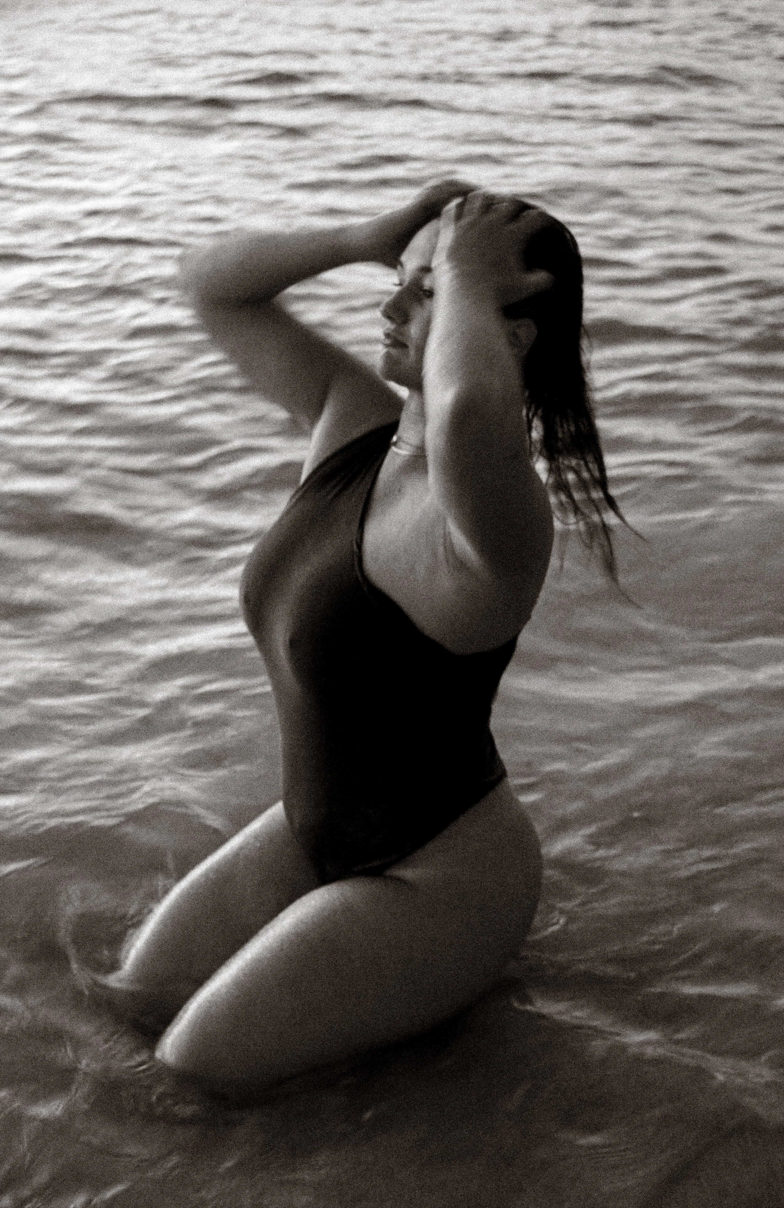 A woman kneeling in water, wearing a swimsuit, with her hands in her hair, eyes closed, and facing sideways.