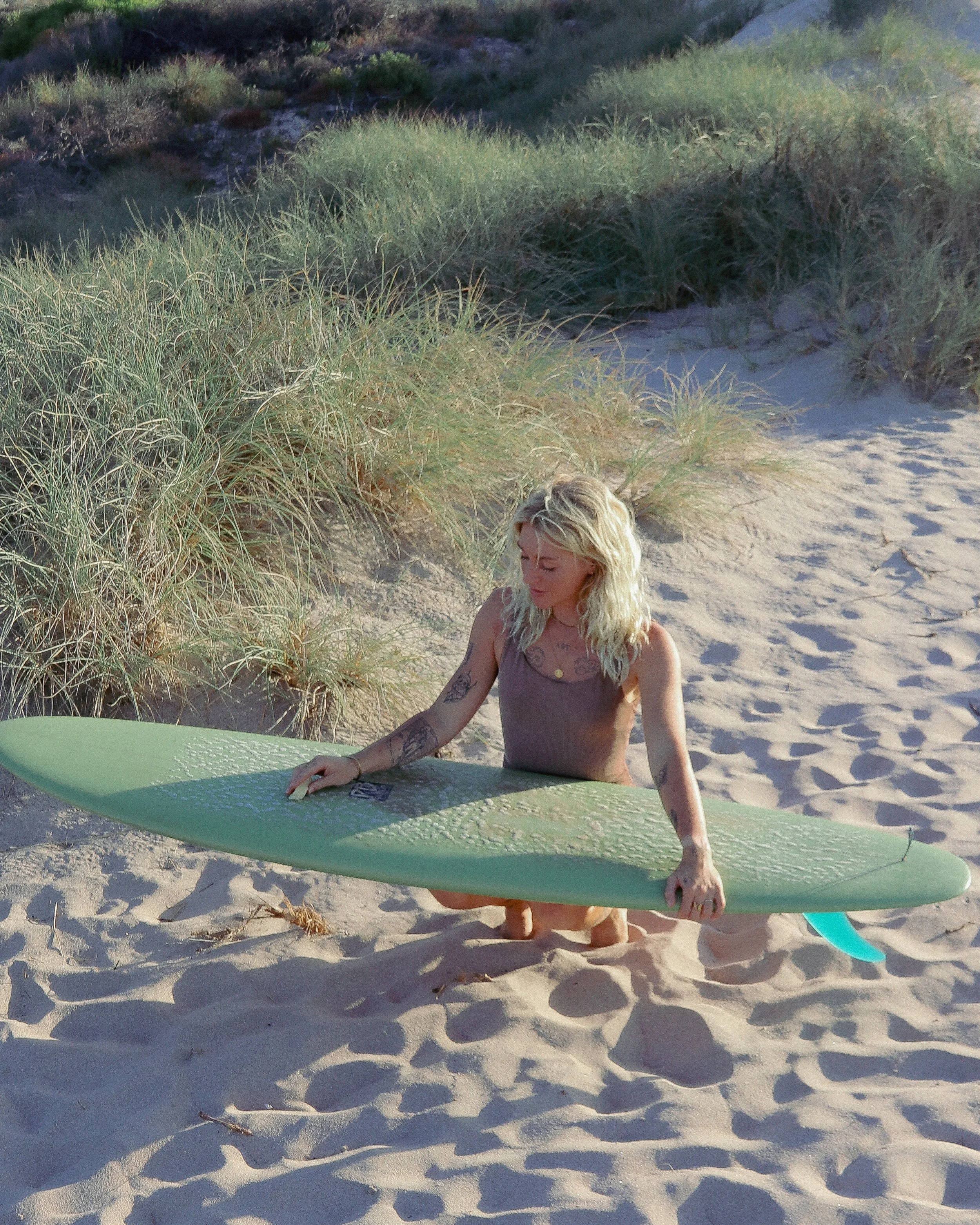 A woman with blonde hair and tattoos kneeling on the sandy beach, holding a green surfboard.