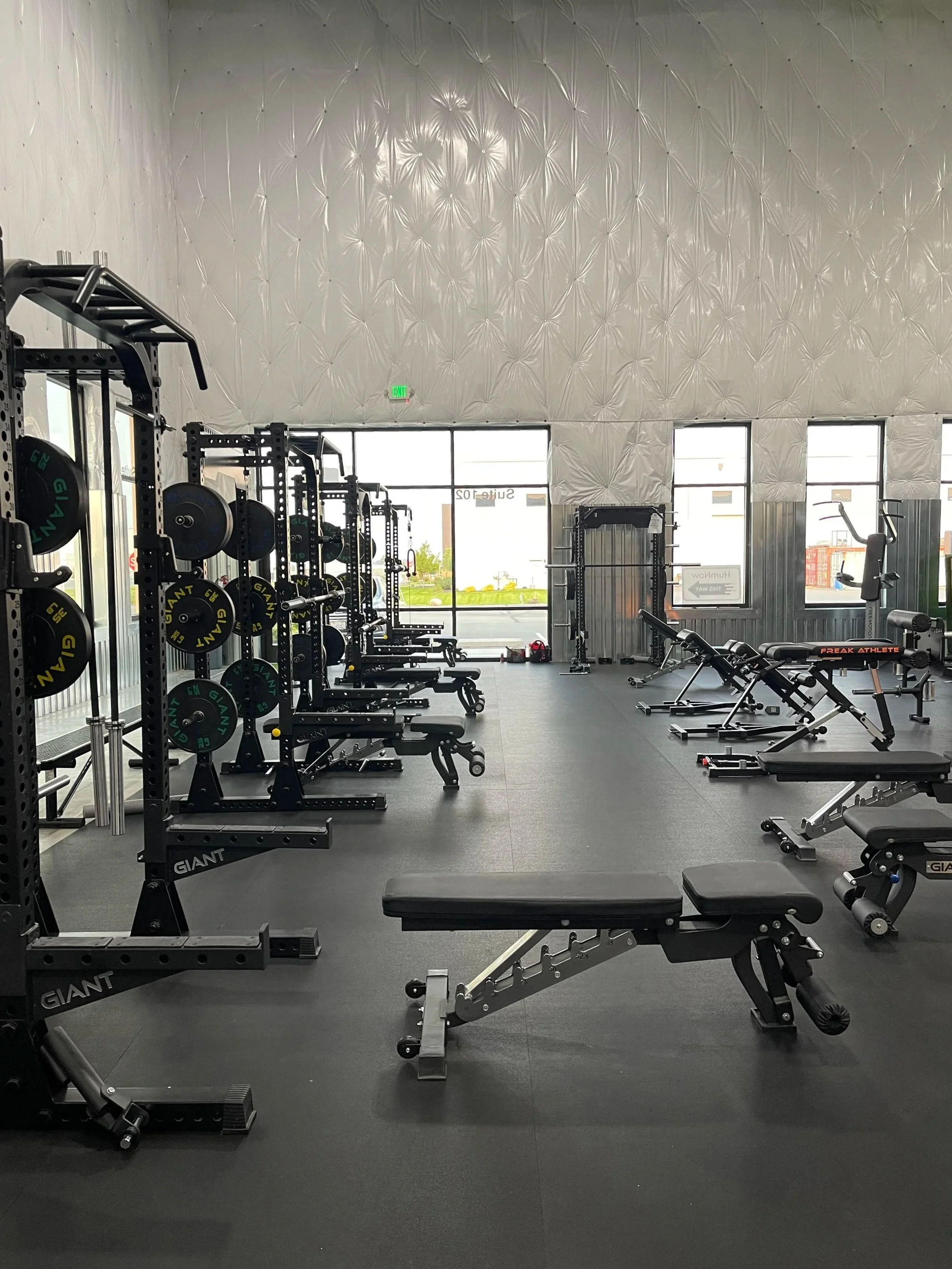 Empty gym with weightlifting equipment, benches, and exercise machines near large windows.