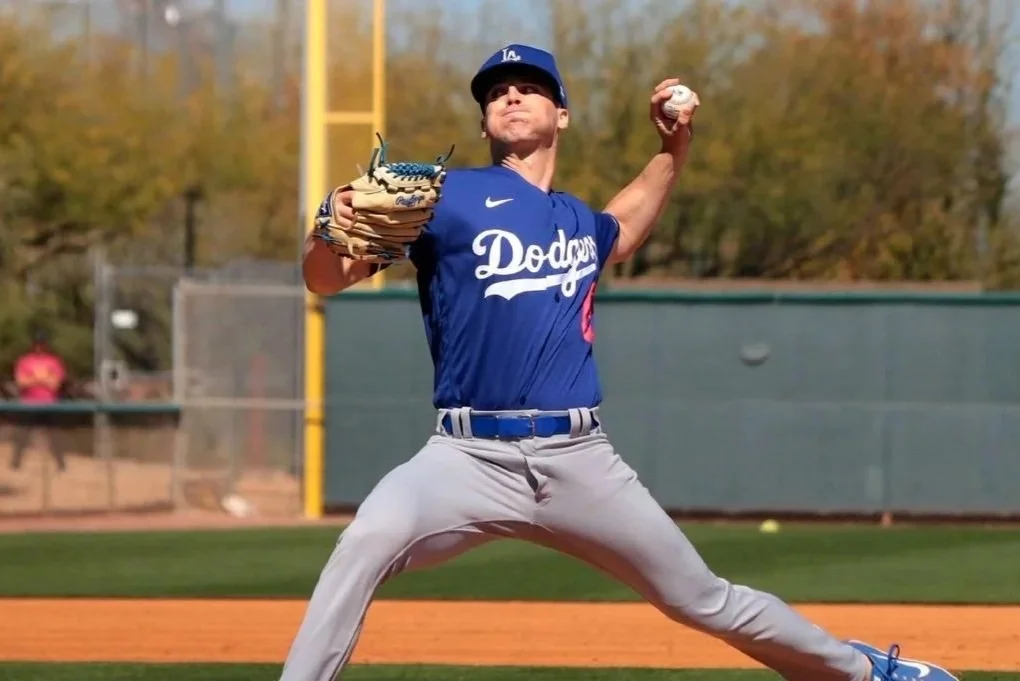 A baseball player in a blue Dodgers jersey and gray pants on the field, preparing to pitch, with a glove in his left hand and a baseball in his right hand, with trees and a fence in the background.