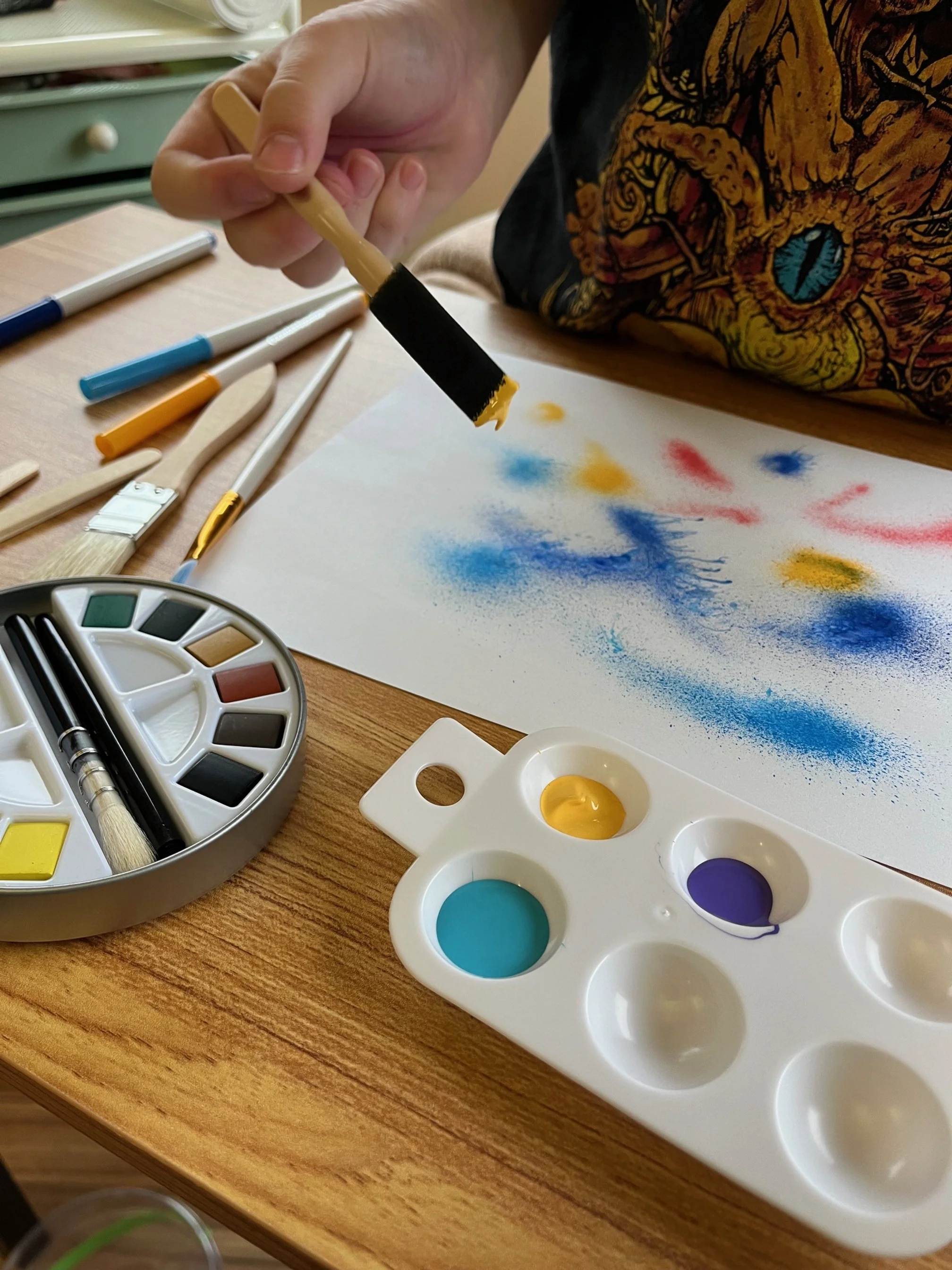A person is using a black and yellow paintbrush to create colorful splashes of blue, yellow, red, and purple on a piece of white paper. Various art supplies, including markers, paintbrushes, and watercolor paints in blue, yellow, and purple, are arranged on a wooden table.