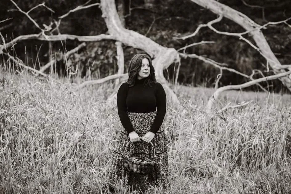 A woman standing in a grassy field holding a basket, with a large, fallen tree in the background.