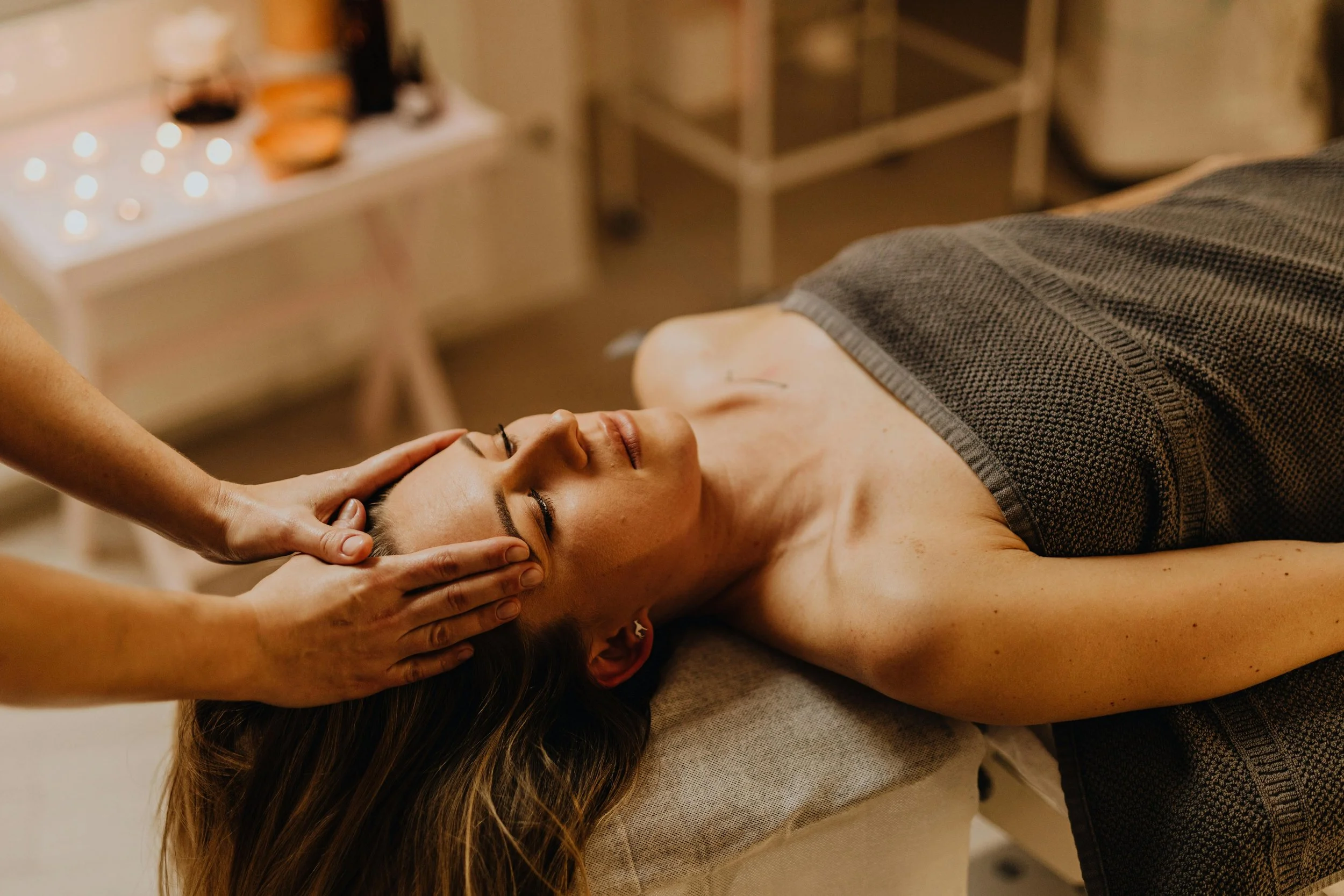 A woman lying on a massage table with a towel covering her body, receiving a massage or facial treatment in a spa or wellness center, with her eyes closed and a serene expression.