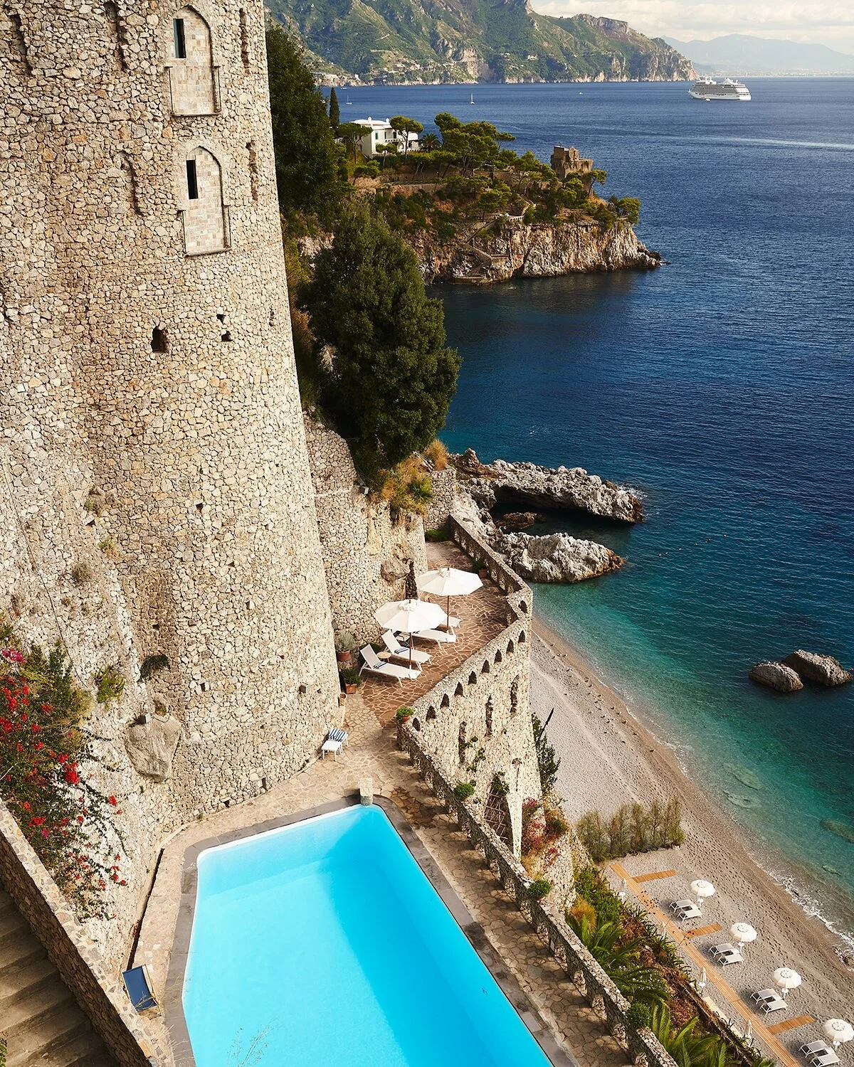 Amalfi Coast - A scenic view of a historic stone building with a terrace, swimming pool, and lounge chairs overlooking a beach and blue ocean with a cruise ship, cliffs, and mountains in the background.