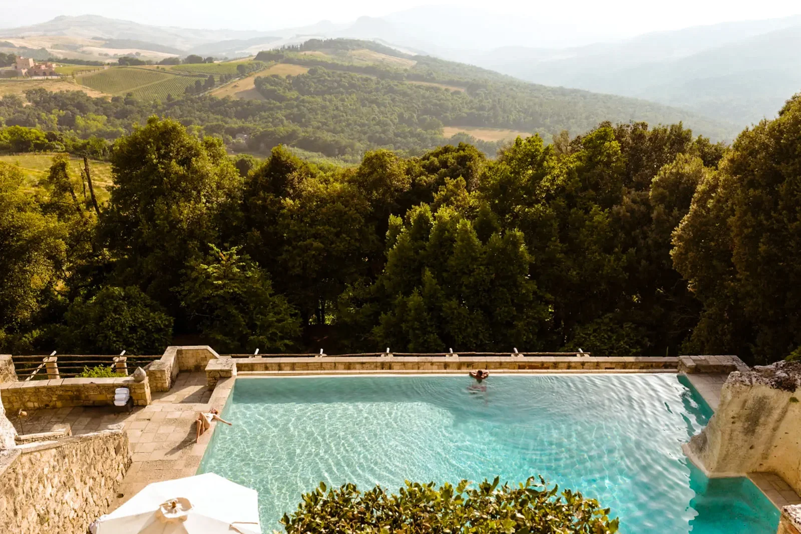A swimming pool in Tuscany with two people relaxing on the edge, overlooking green hills and trees in a scenic countryside landscape.