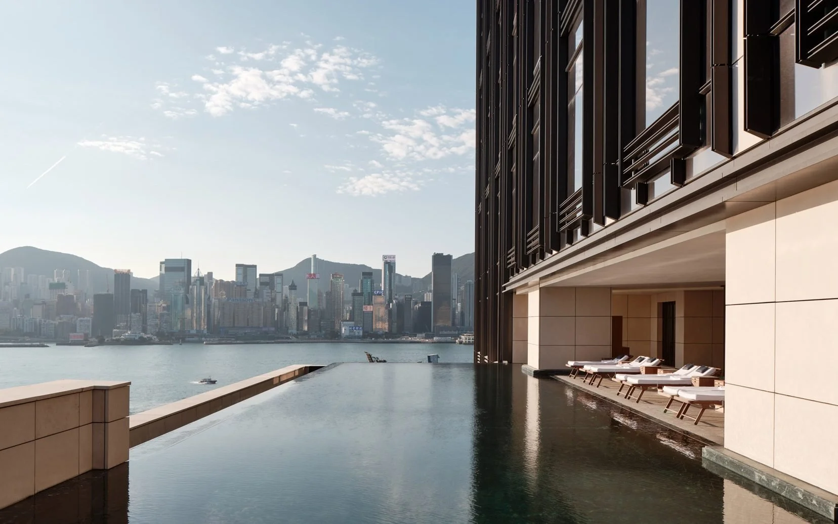 Hong Kong hotel with sun loungers beside an infinity pool overlooking a city skyline, harbor, and mountains, on a clear day.