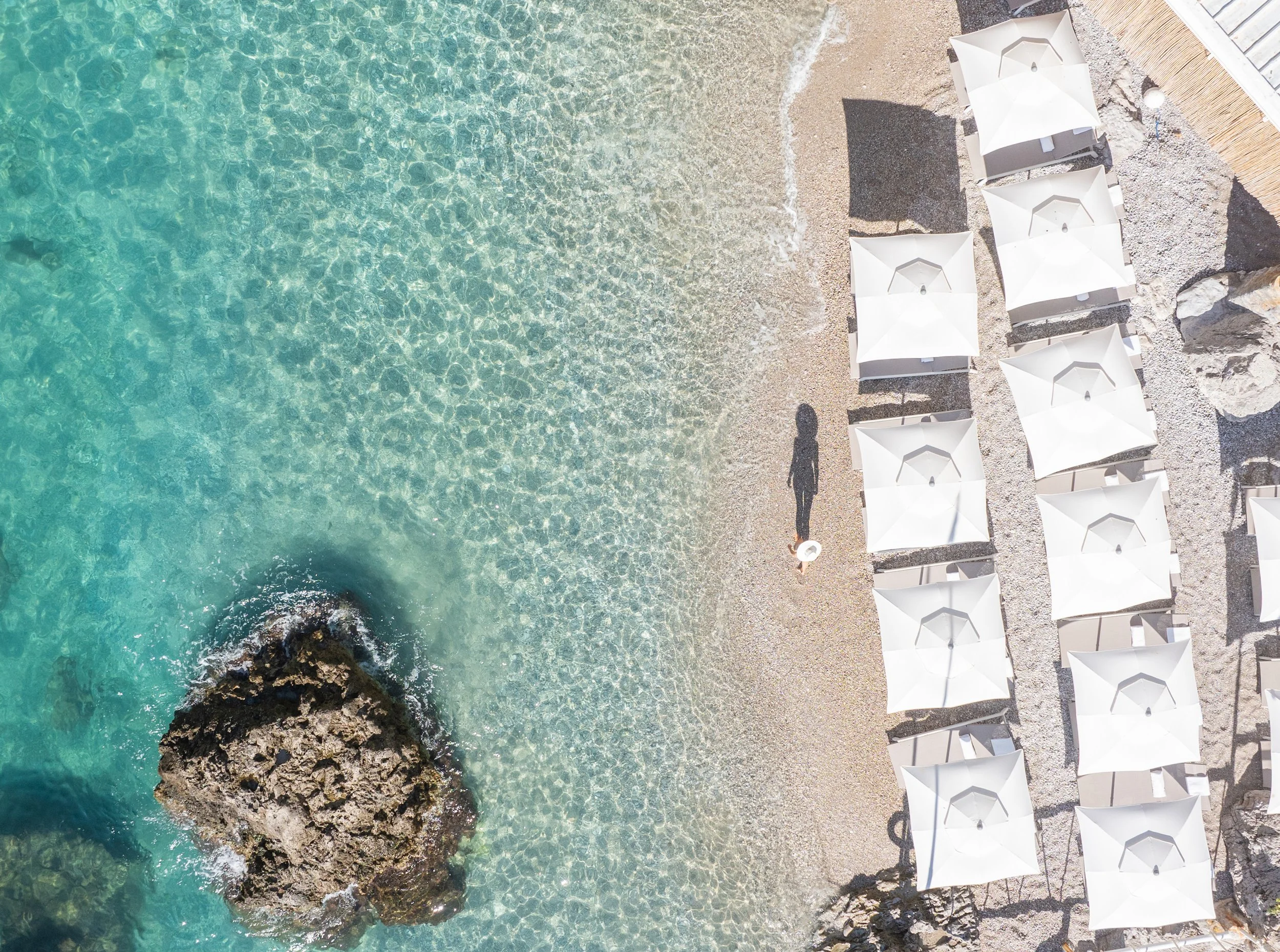 Capri, Italy - Aerial view of a beach with clear turquoise water, a large rock near the shoreline, and a row of white beach umbrellas on sand. A person is walking along the shore carrying a hat.