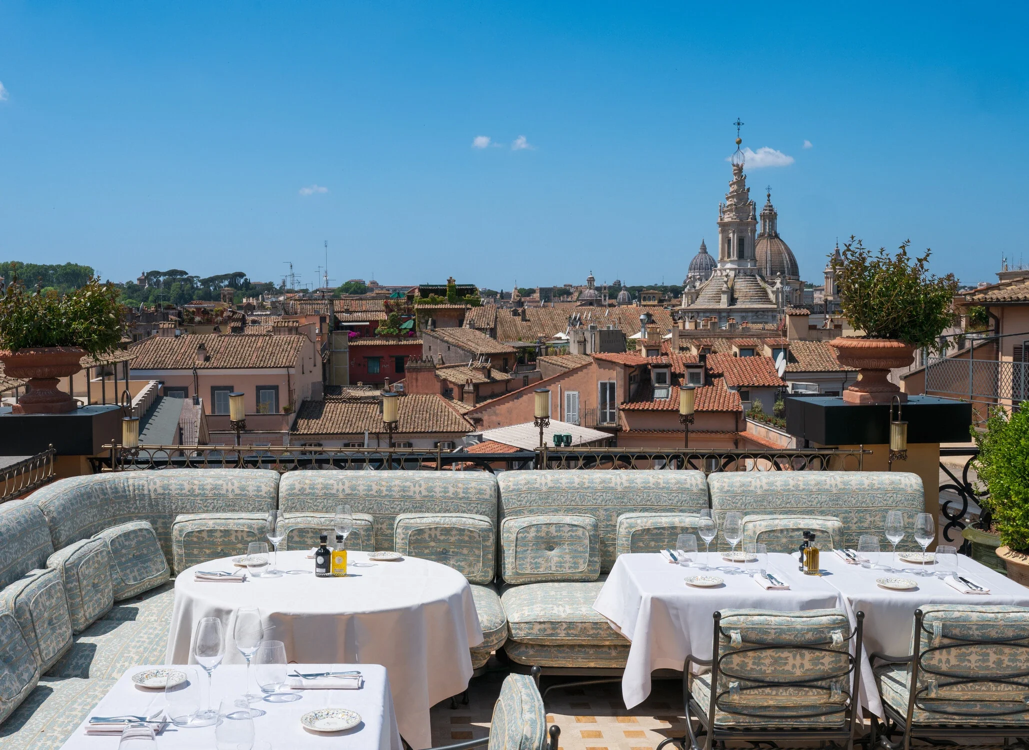 Rome, Orient Express La Minerva - Outdoor restaurant terrace with white tablecloths, glassware, and cutlery, overlooking a cityscape with terracotta rooftops and historic buildings under a blue sky.