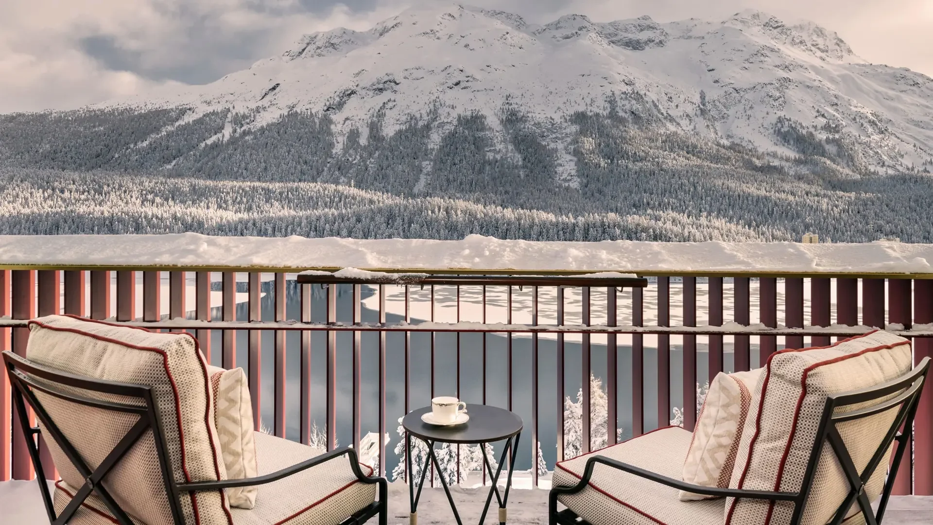St Moritz - A snowy mountain view from a balcony with two chairs and a small table with a cup. Snow covers the railing and balcony floor, with a mountain, snowy forest, and cloudy sky in the background.