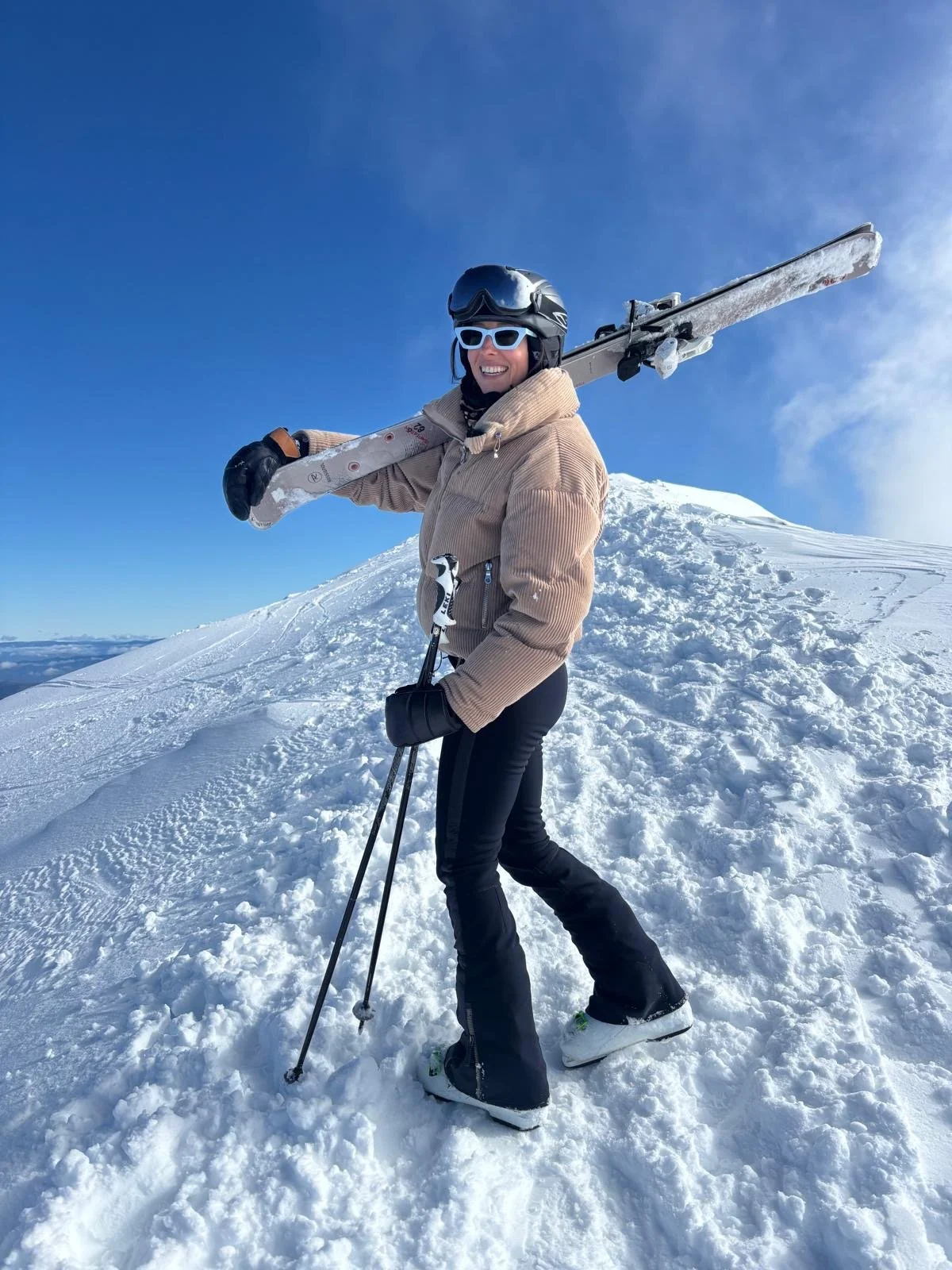 Woman in winter gear standing on snowy slope, holding a pair of skis over her shoulder, smiling, wearing helmet and goggles, blue sky in the background.