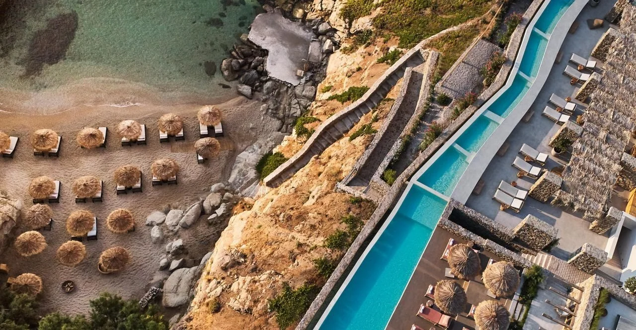 Aerial view of a resort in Mykonos with a long infinity pool, sun loungers, and thatched umbrellas.