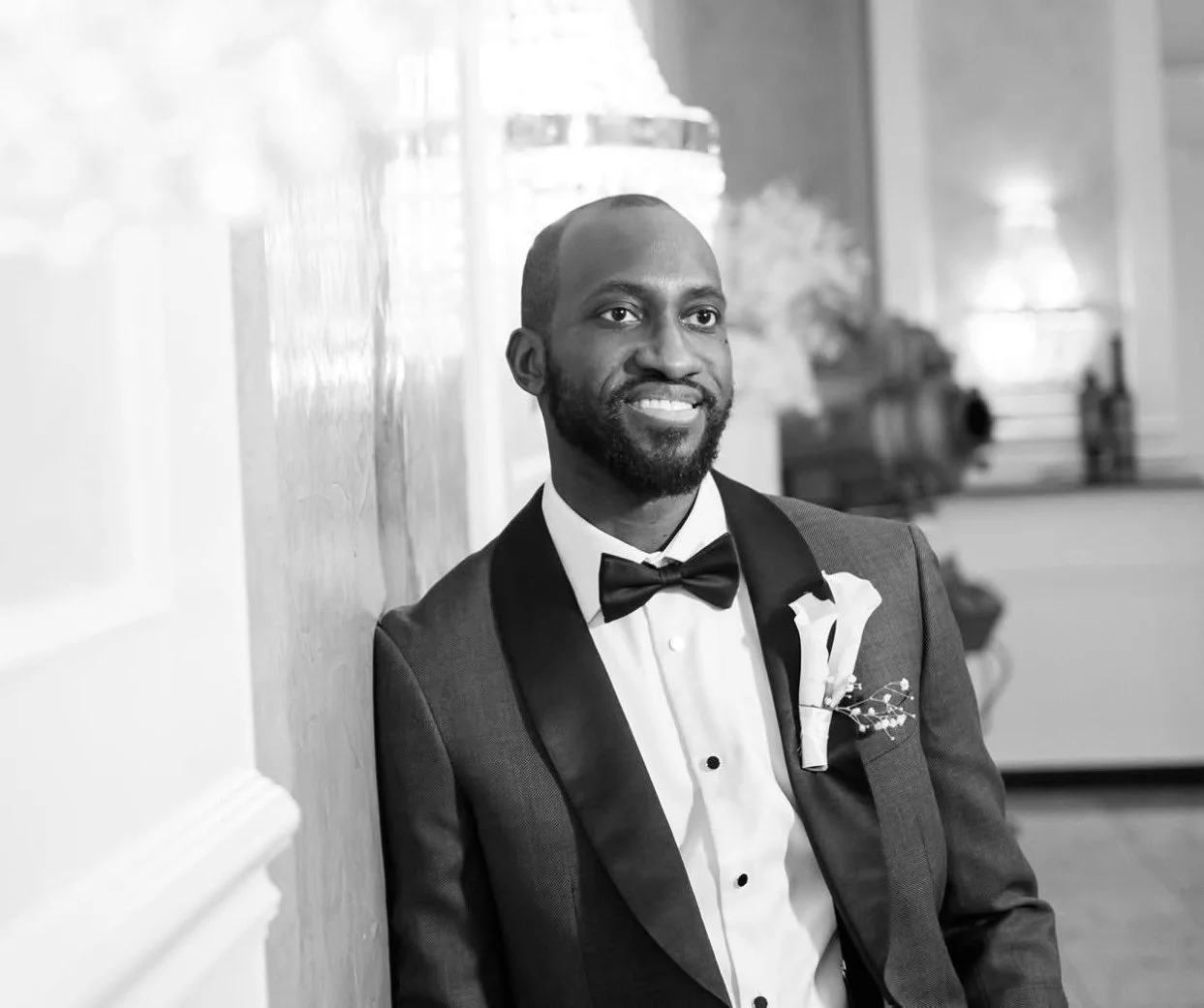 A man in a tuxedo with a bow tie and boutonniere, smiling while leaning against a wall in an elegant room.