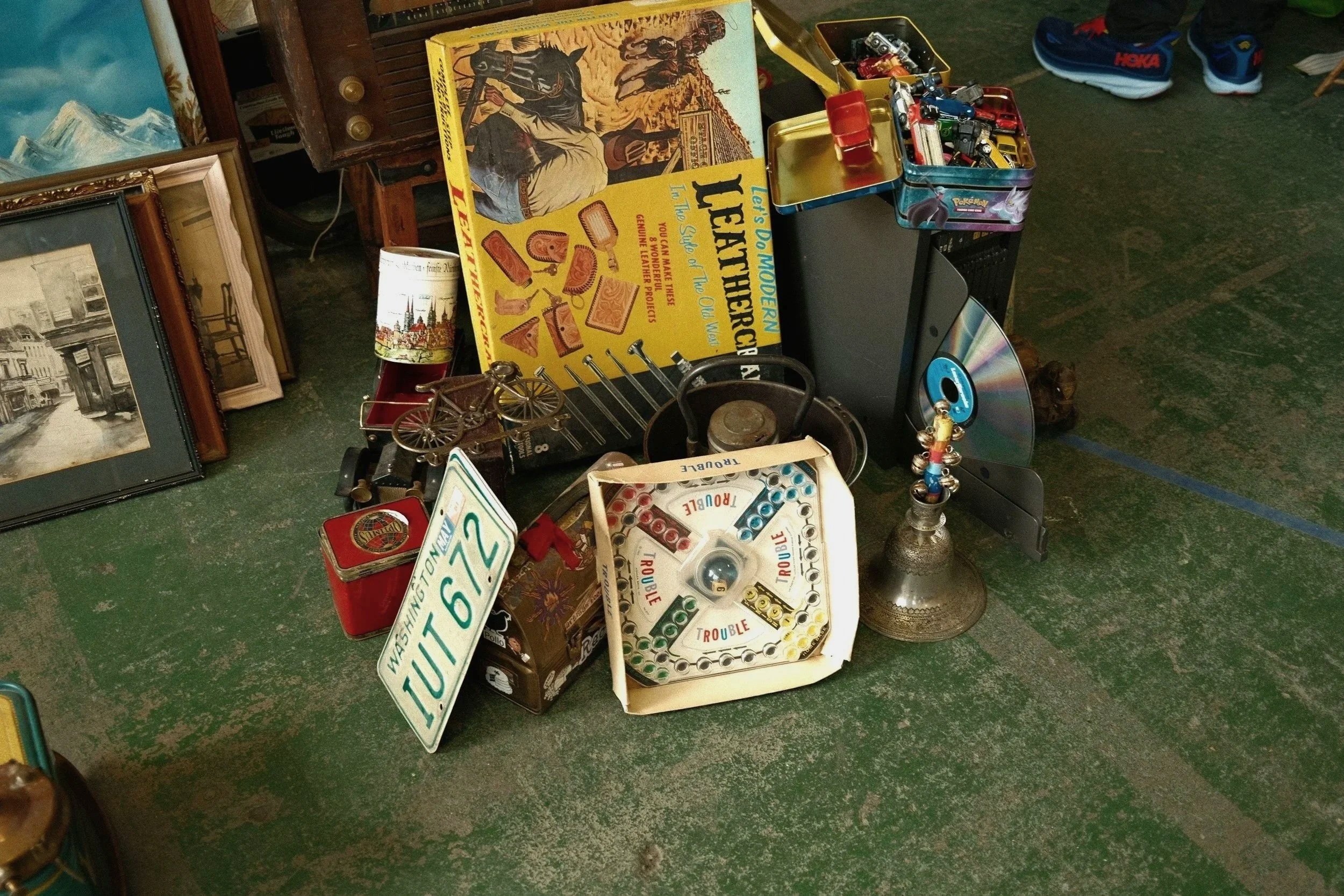 A collection of vintage and collectible items, including a vintage toy car, a board game called 'Let's Do Modern Leaf Herb Craft,' framed pictures, an old bicycle model, a license plate from Washington, a speaker, a CD, and various other small items on a green floor.