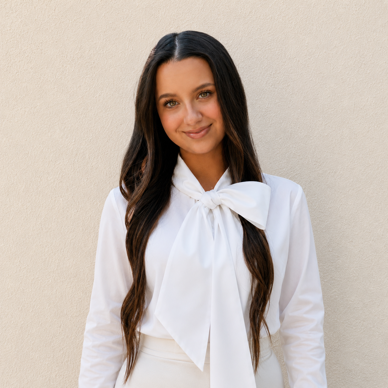 Young woman with long dark hair wearing a white blouse with a bow tie, standing against a neutral wall, smiling softly.
