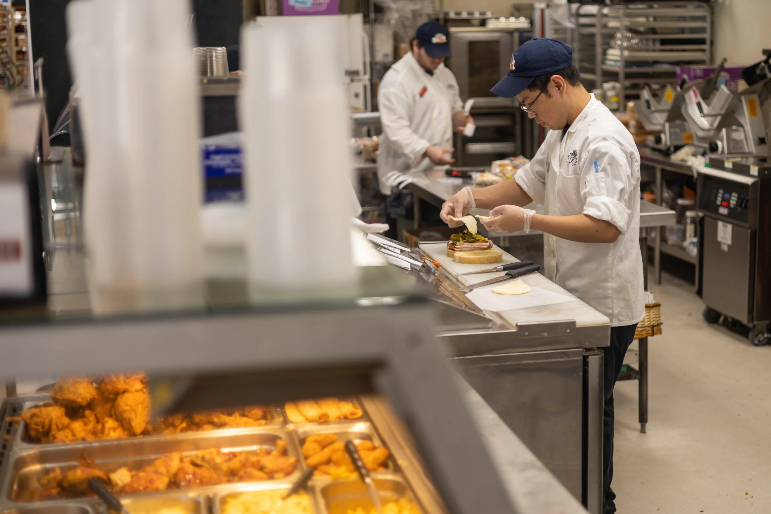Two kitchen workers preparing sandwiches in a commercial kitchen with trays of fried chicken and other food items.