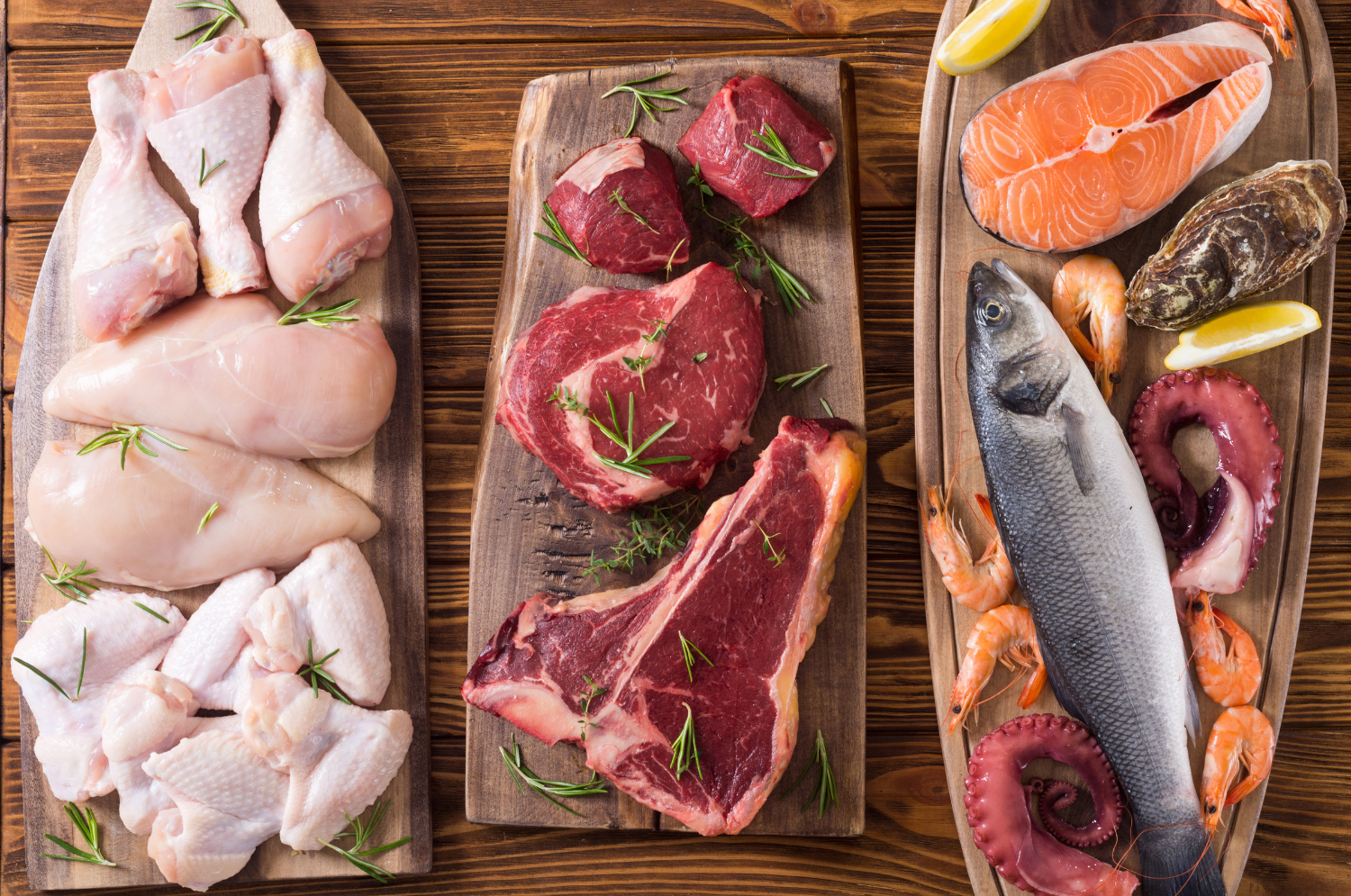 Three wooden boards with raw chicken pieces, beef steaks, and assorted seafood including salmon, shrimp, octopus, and oysters, garnished with lemon wedges and herbs on a wooden table.