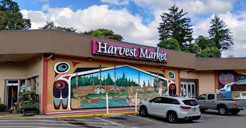 Harvest Market grocery store with colorful mural of a theater scene, parked cars in front, and trees in the background.