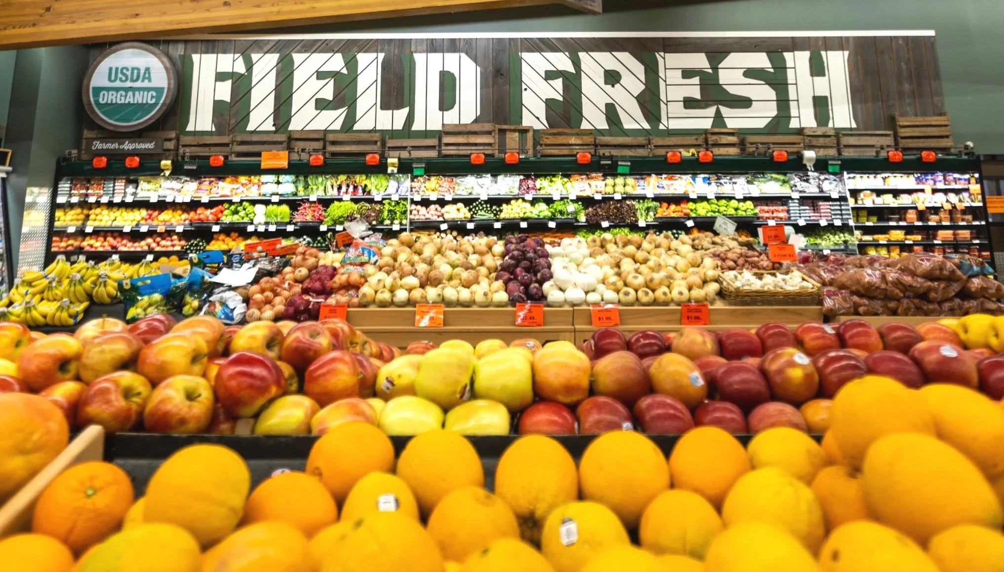 Fresh fruits and vegetables display at a grocery store produce section, with apples, bananas, onions, and other produce arranged in bins and shelves.