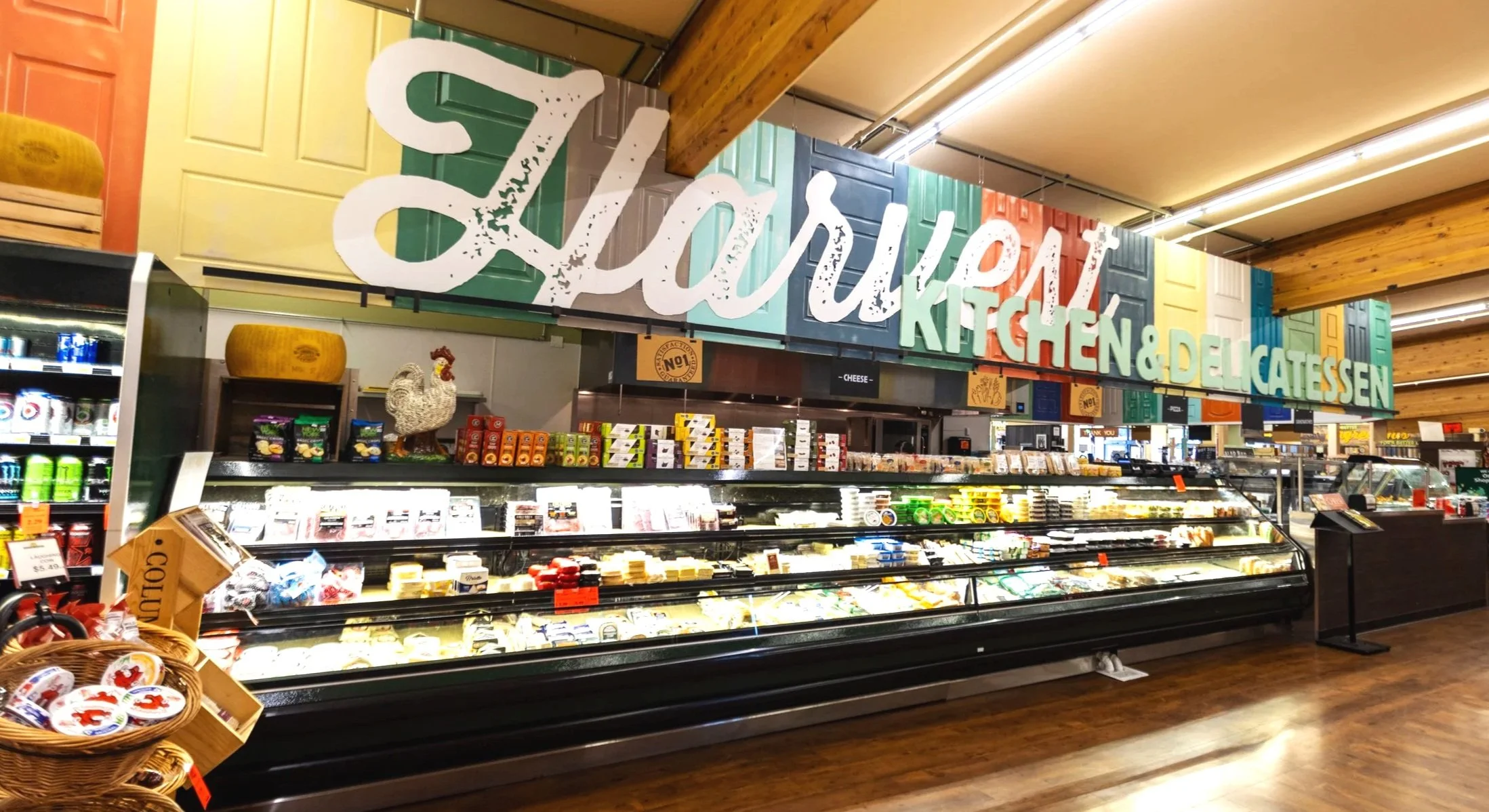 Deli section in a grocery store with cheese and delicatessen products behind glass, signage overhead reading 'Salumi Kitchen & Delicatessen', and a basket of pre-packaged cheese or spreads in the foreground.