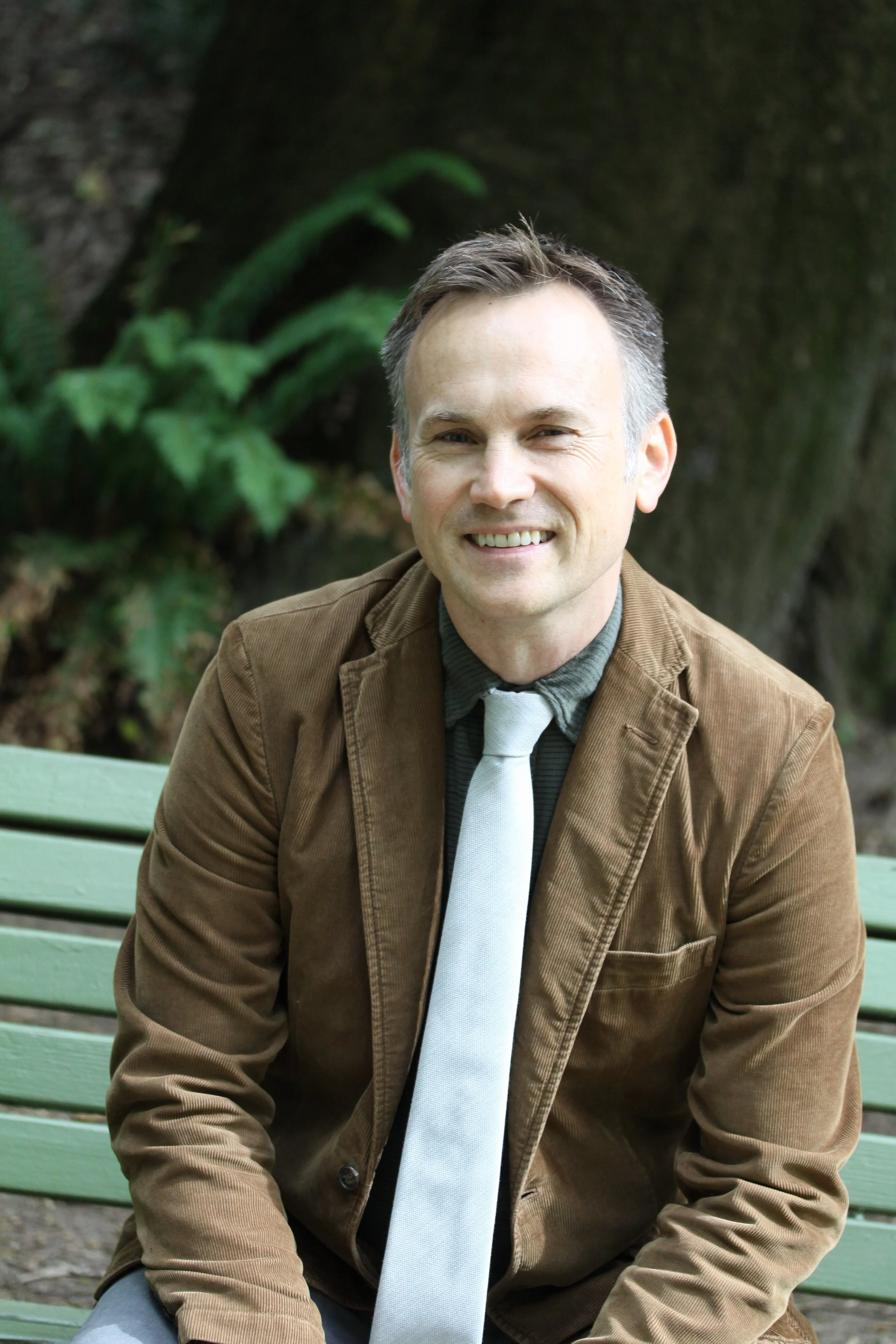 A middle-aged man with gray hair, smiling and sitting on a green park bench outdoors, wearing a brown jacket, gray shirt, and light gray tie, with trees and greenery in the background.