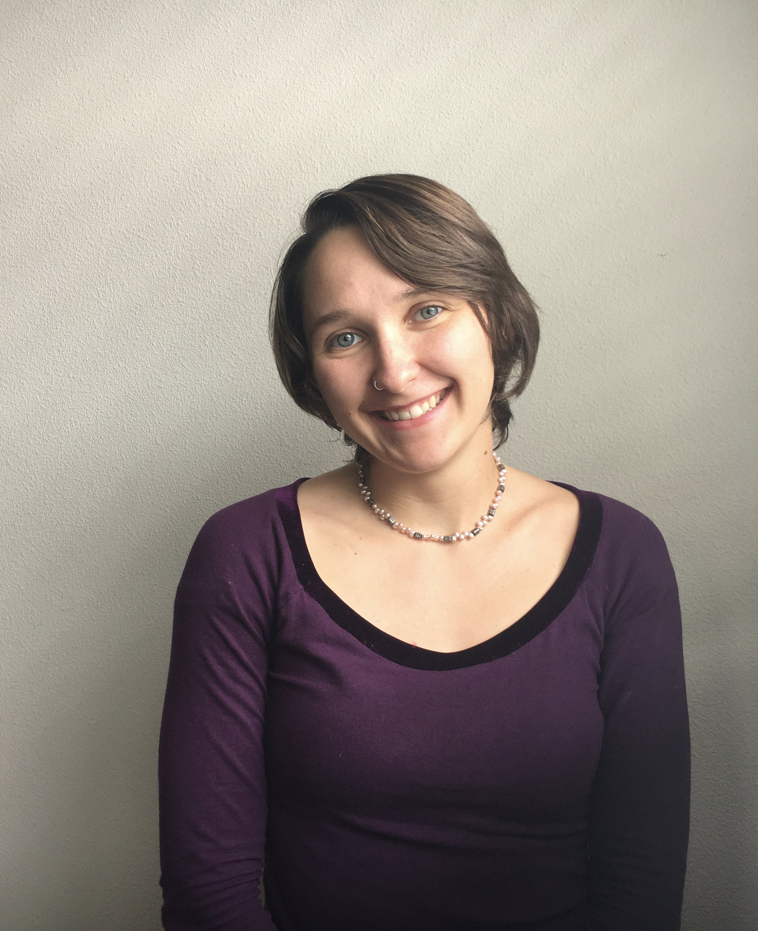 A young woman with short brown hair, blue eyes, and a nose ring, smiling at the camera, wearing a purple top and a beaded necklace, standing against a plain light-colored wall.