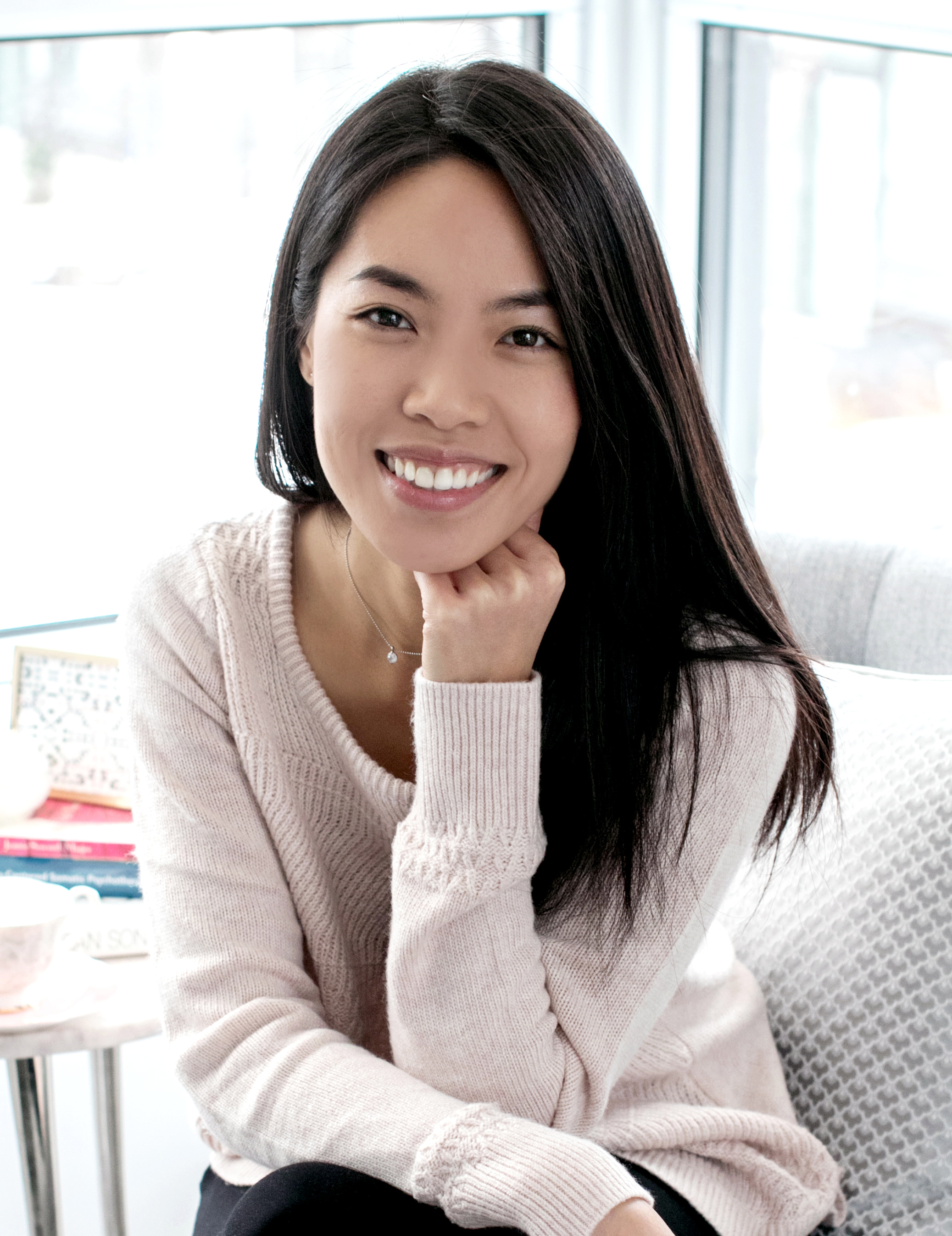 A woman with long dark hair smiling, sitting indoors near large windows, wearing a light pink sweater.