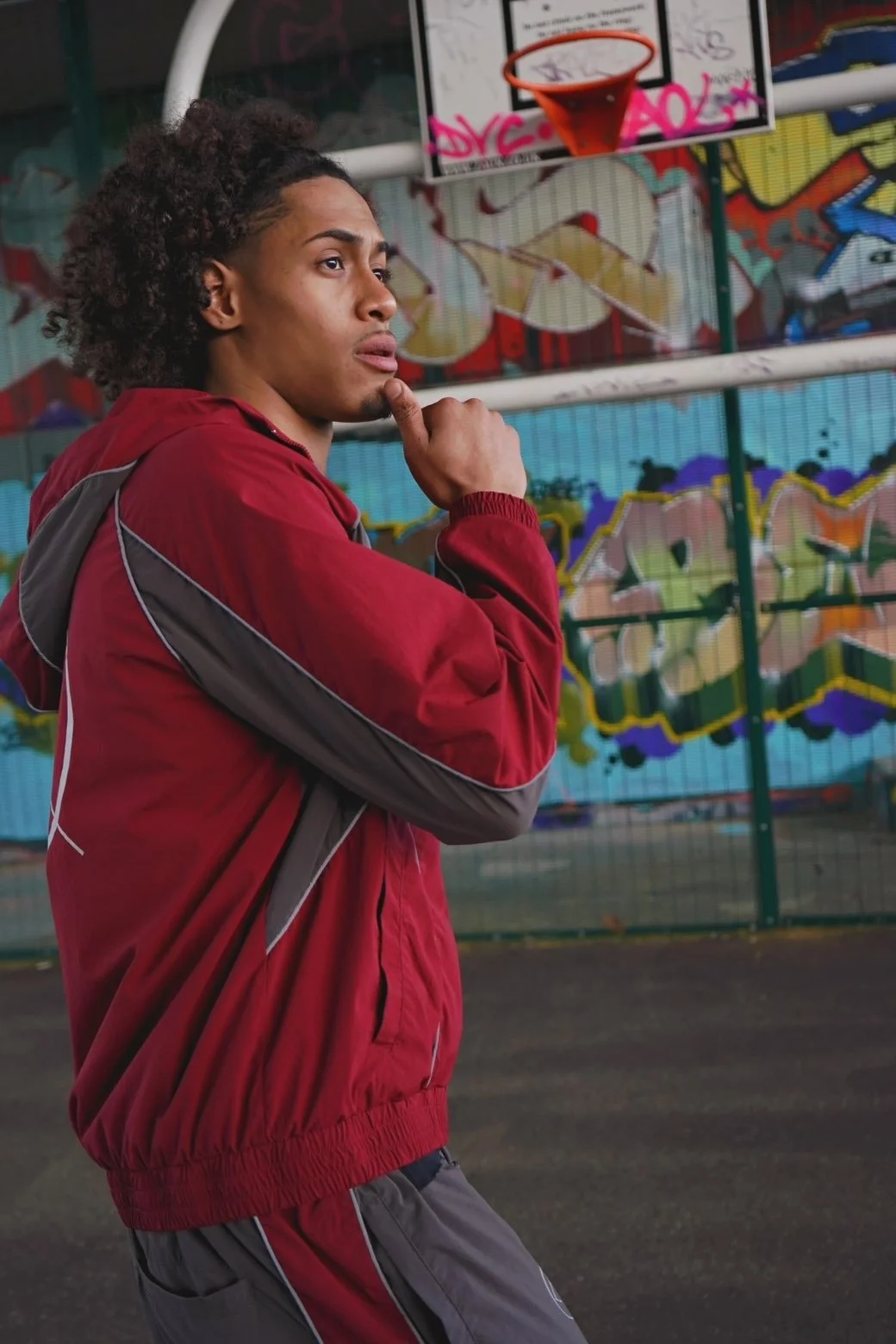 A young man in a red and gray jacket standing near a colorful graffiti-covered wall and a basketball hoop, looking contemplative.