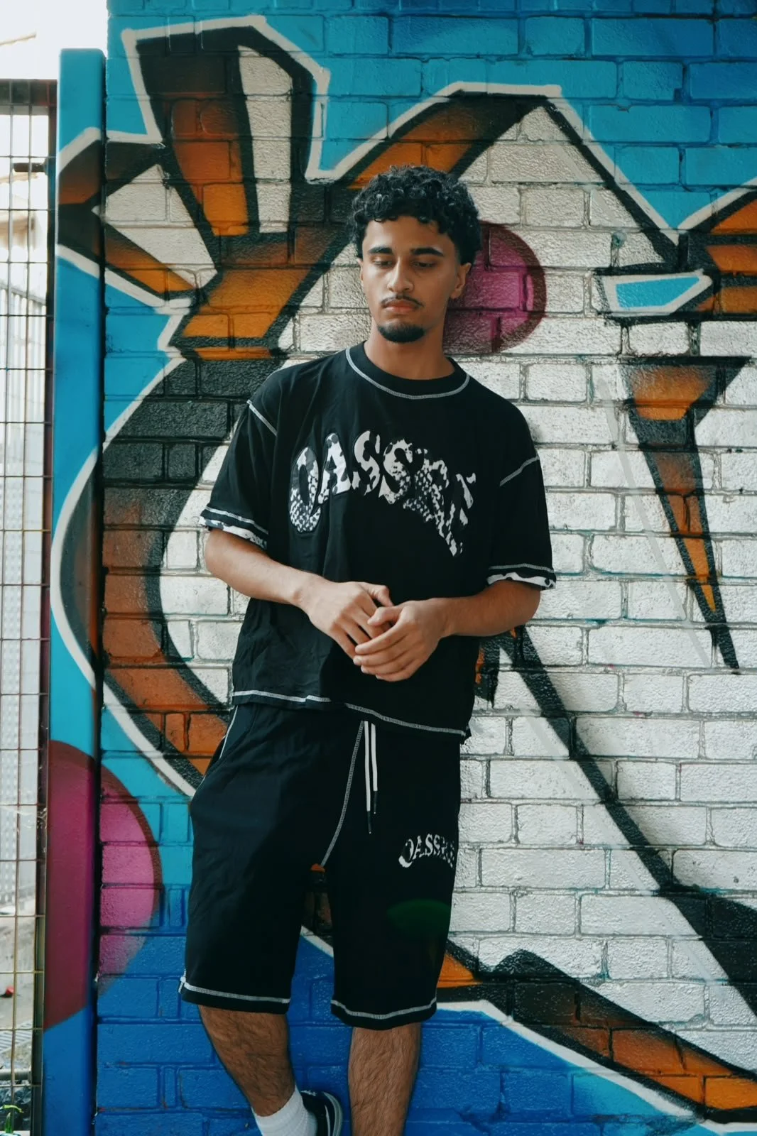 A young man with curly black hair, wearing a black t-shirt and shorts, standing against a colorful graffiti mural on a brick wall.