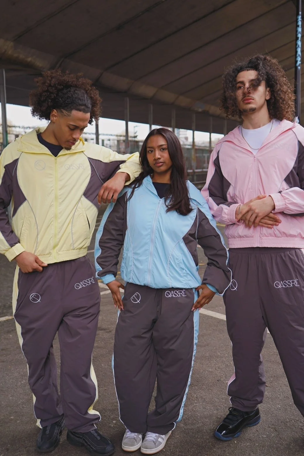Three young people wearing sporty track jackets and pants under an overpass, looking serious. The person on the left is a man with curly hair in a yellow and gray tracksuit, the middle person is a woman with straight hair in a light blue and gray tracksuit, and the person on the right is a man with curly hair in a pink and gray tracksuit.