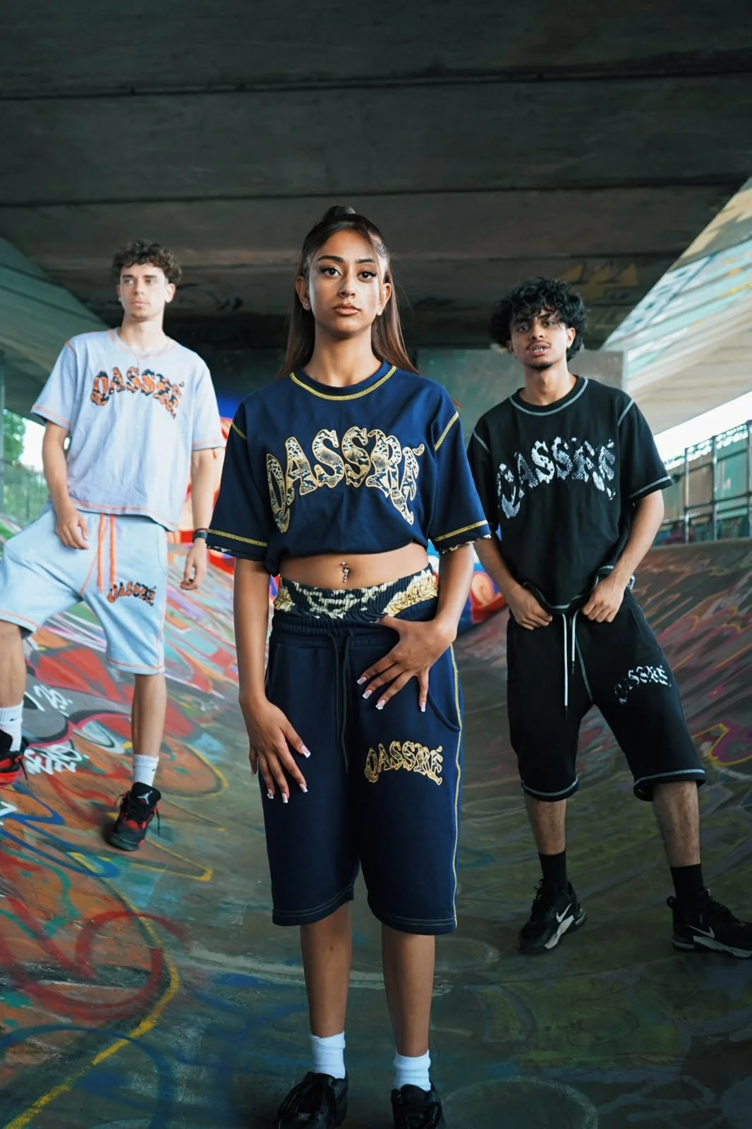 Three young people standing in a graffiti-covered skate park, wearing matching sporty outfits with the word 'OASSSS' written on their clothing.