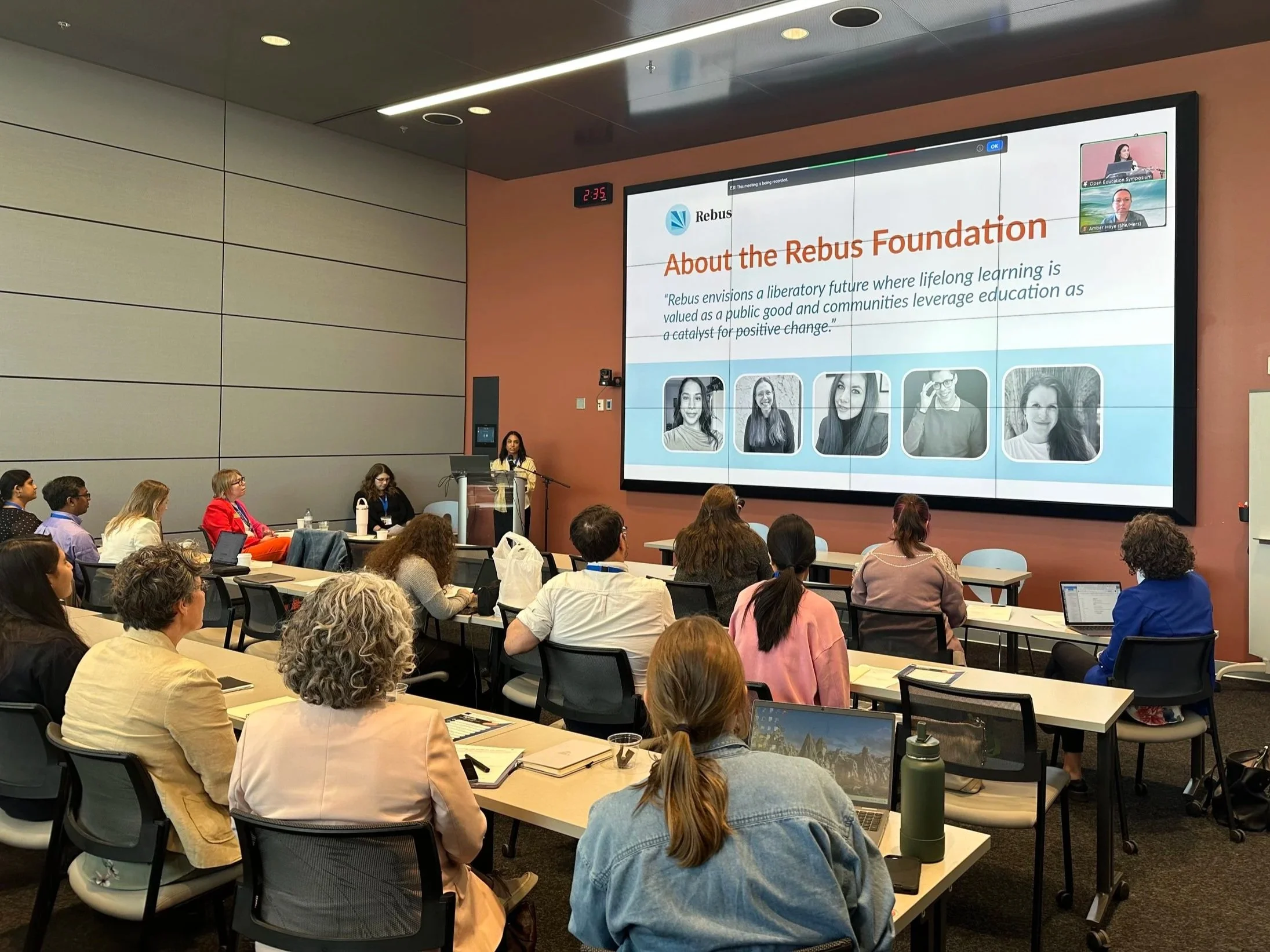 A diverse group of people attending a conference or presentation in a modern auditorium. A large screen displays a slide titled 'About the Rebus Foundation' with images of people, and a woman is speaking at a podium.