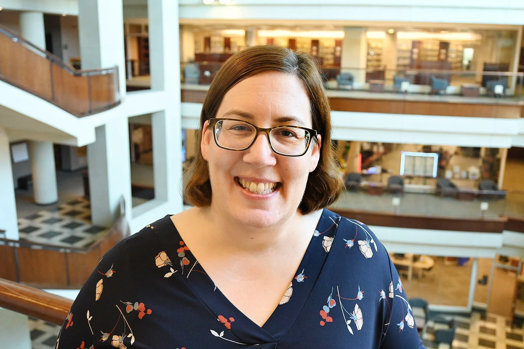 Ginelle faces the camera, wearing a floral blouse and glasses. In the background is a multi-level library.