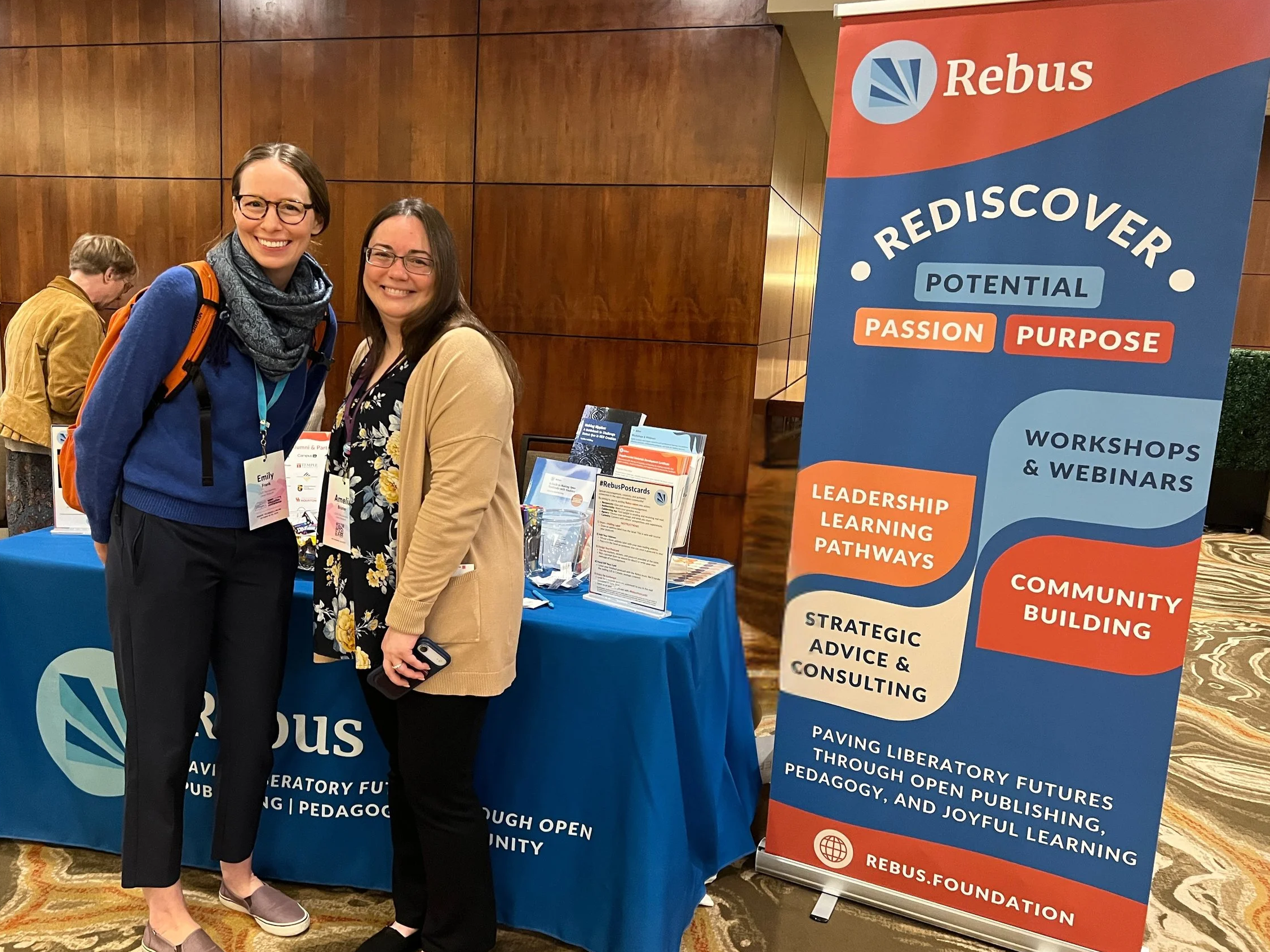 Two women standing next to a Rebus Foundation banner at a conference or event, smiling at the camera.