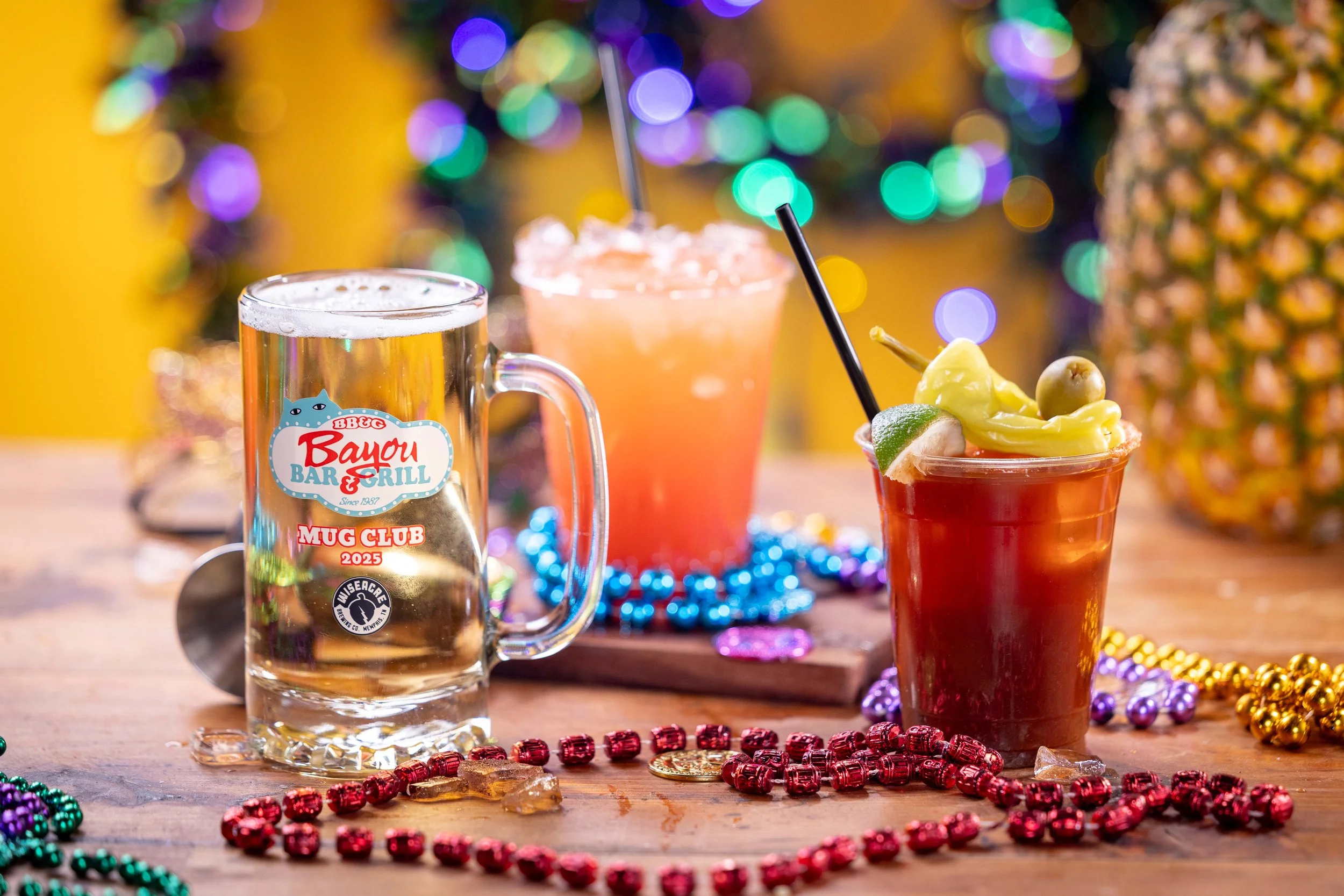 Three colorful cocktails on a wooden table with Mardi Gras beads, a pineapple, and blurred festive lights in the background.