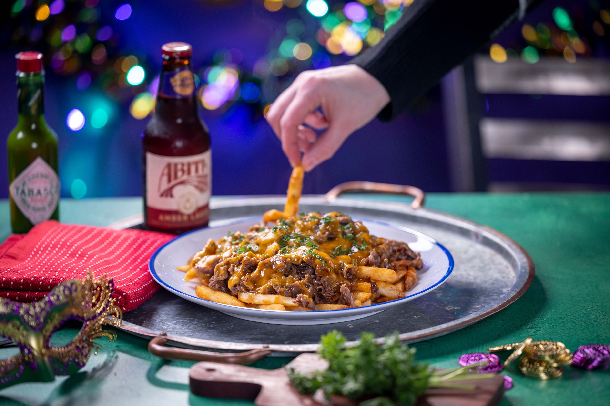 Person sprinkling cheese on a plate of loaded fries with ground beef, cheese, and chopped green herbs, served on a silver tray. Two bottles of beer and Mardi Gras decorations are on a green tablecloth, with colorful blurred lights in the background.
