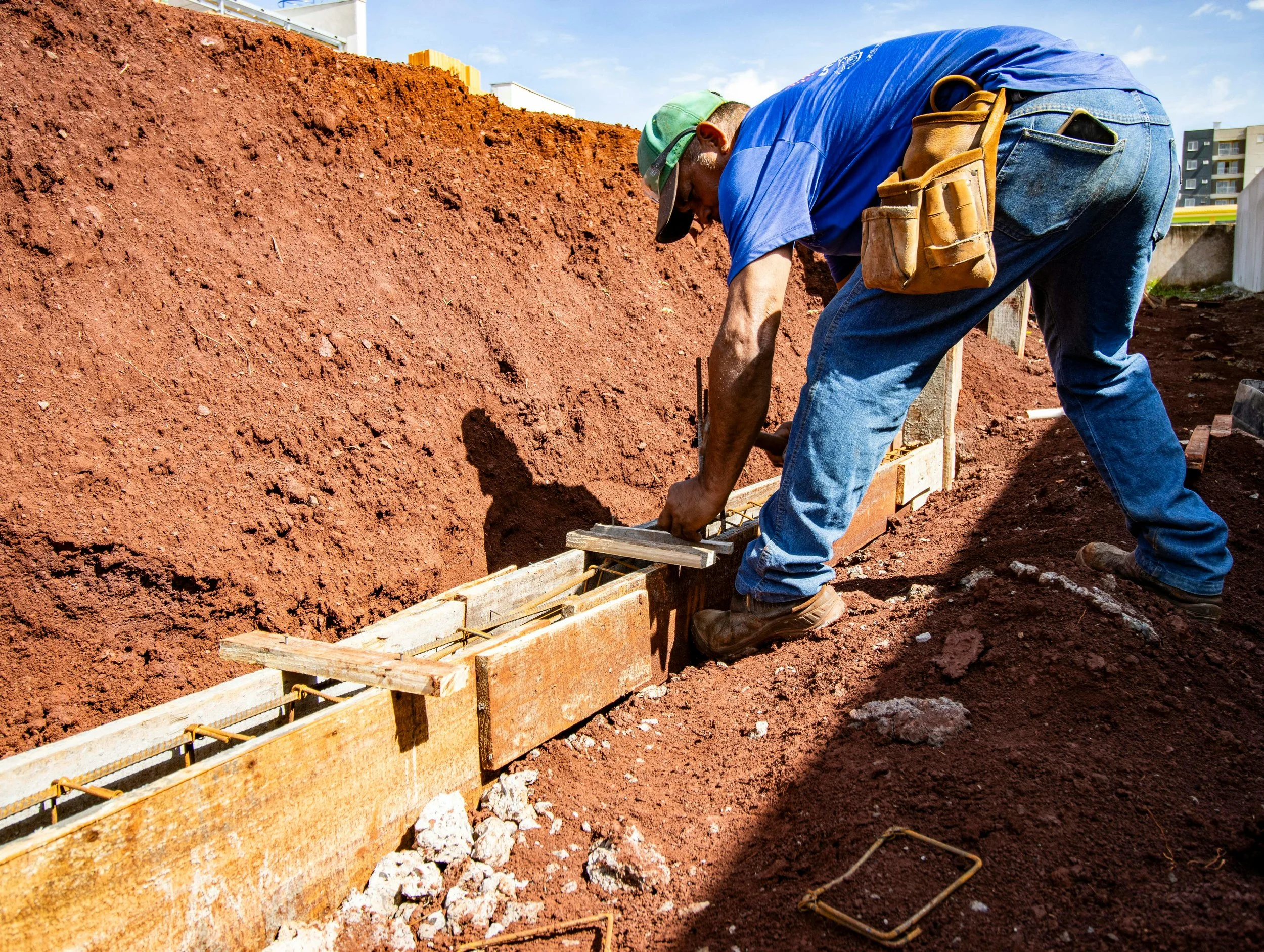 A construction worker in a blue shirt, jeans, and a cap is working on building a foundation on a red dirt site. The worker is bending over, securing wooden formwork for concrete.