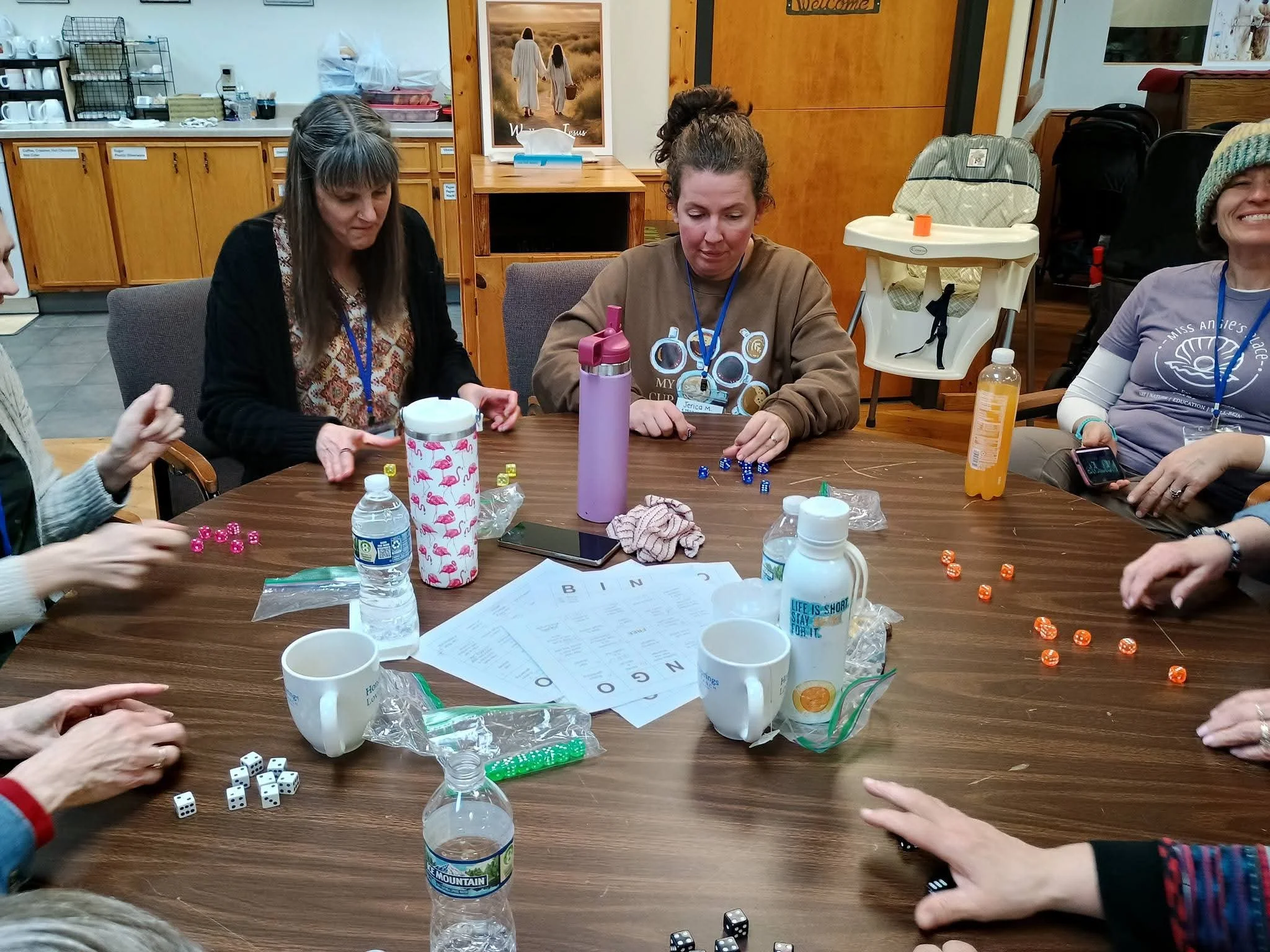 A group of women sitting at a wooden table, playing a game involving dice, with cups, water bottles, and small game pieces on the table, in a room with wooden cabinets, posters, and a high chair in the background.