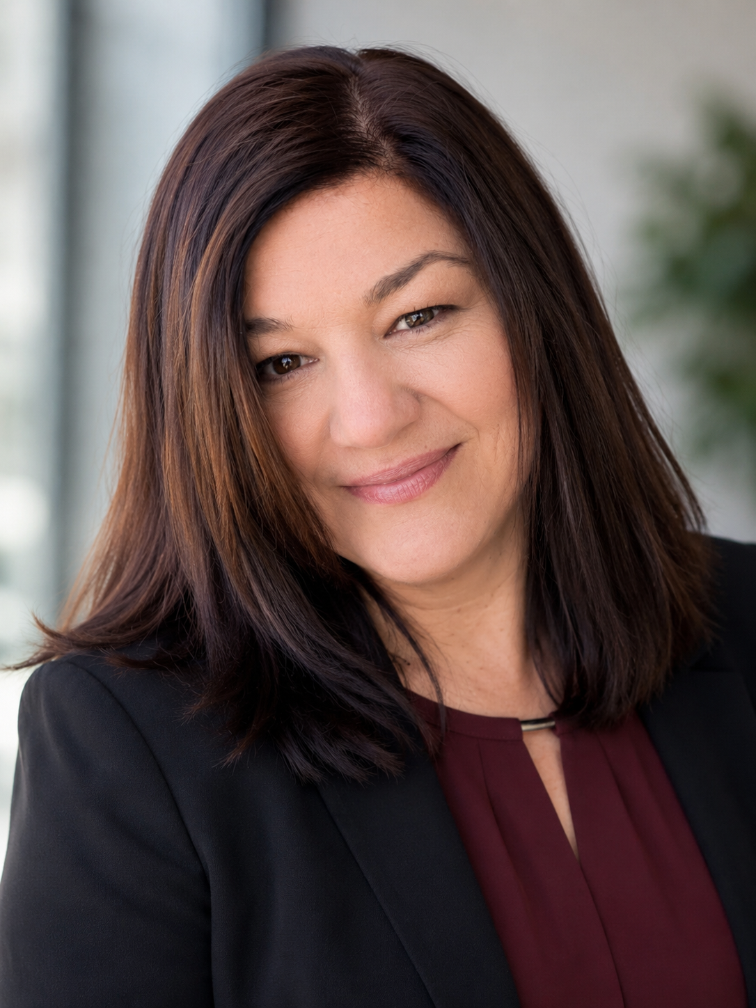 Close-up of a woman with shoulder-length dark brown hair, smiling, wearing a black blazer and a maroon blouse, in a professional indoor setting.