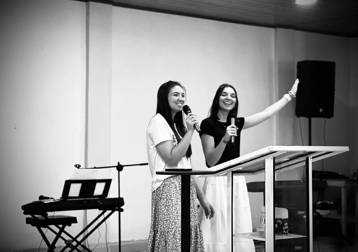 Two young women standing behind a podium, each holding a microphone and smiling, in a room with musical equipment including a keyboard and speaker.