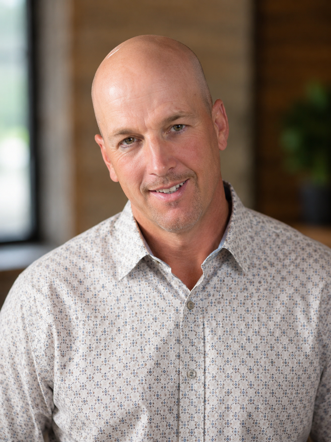 A middle-aged man with a shaved head and light facial hair, wearing a patterned button-up shirt, smiling slightly, in an indoor setting with blurred background.