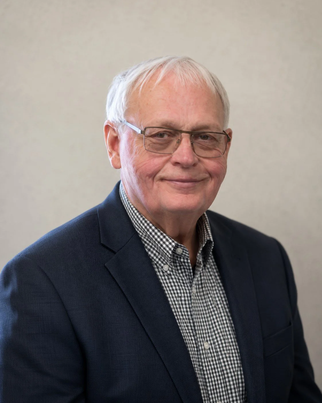 A smiling older man with white hair and glasses wearing a dark blazer and a checkered shirt against a neutral background.