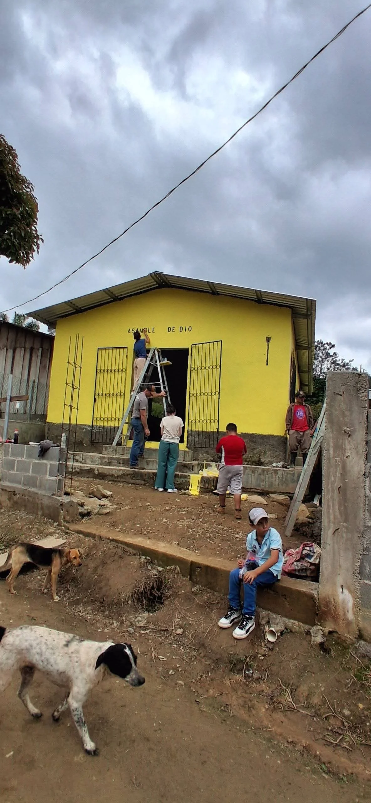 People working on the exterior of a yellow church building with the sign 'Asamblea de Dios', some children and dogs in the foreground, overcast sky.