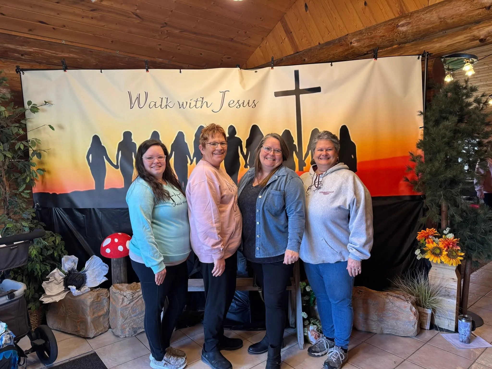 Four women standing close together inside, smiling at the camera, in front of a large banner with silhouettes of people holding hands and the words 'Walk with Jesus.' The background features a cross and sunset colors. Surrounding the group are plants
