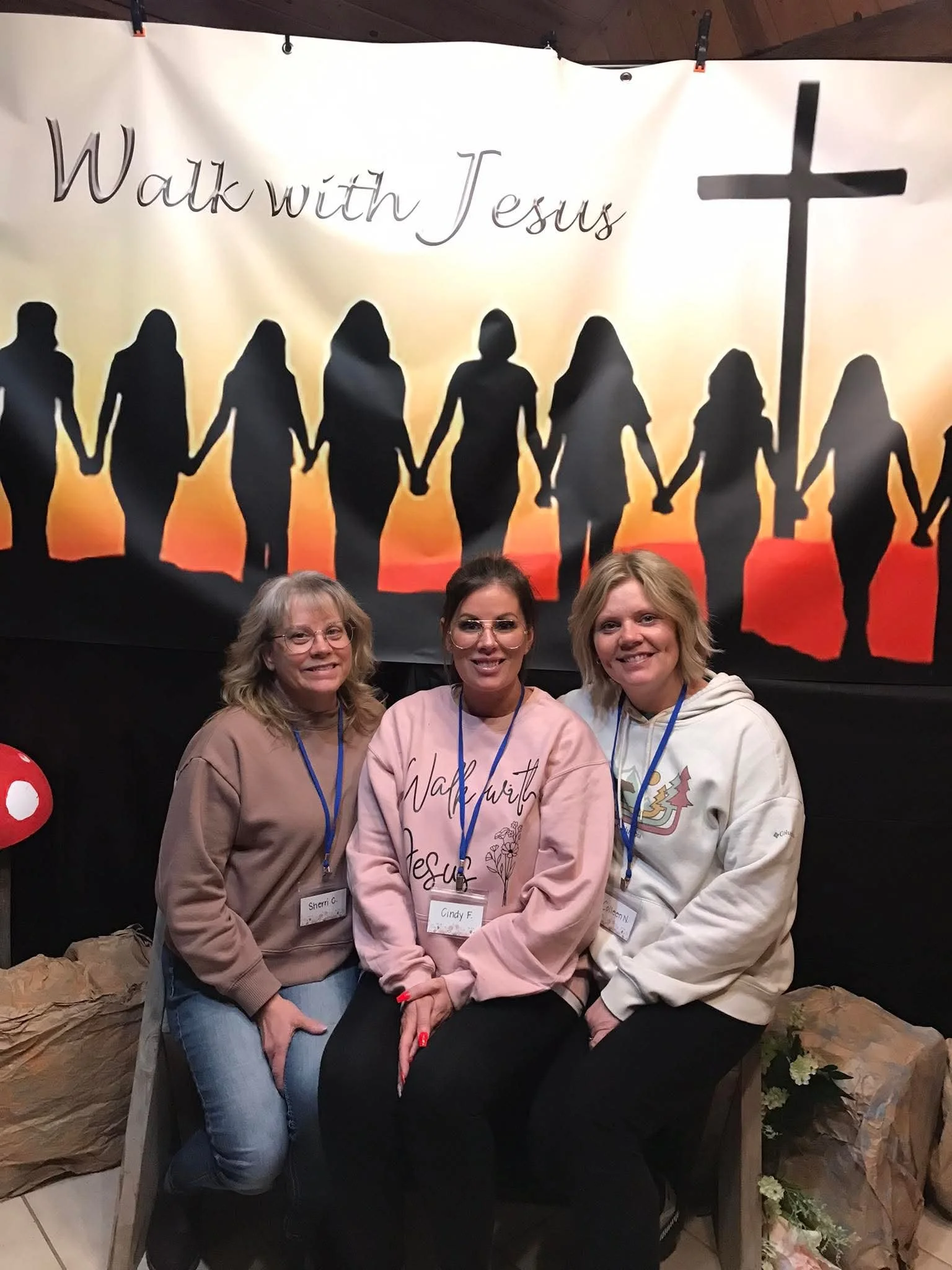 Three women sitting together, smiling in front of a large backdrop with a silhouette of people holding hands and the words 'Walk with Jesus' and a cross