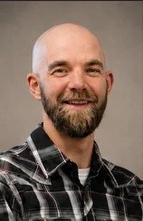 Photograph of a smiling man with a beard and bald head, wearing a checkered shirt.