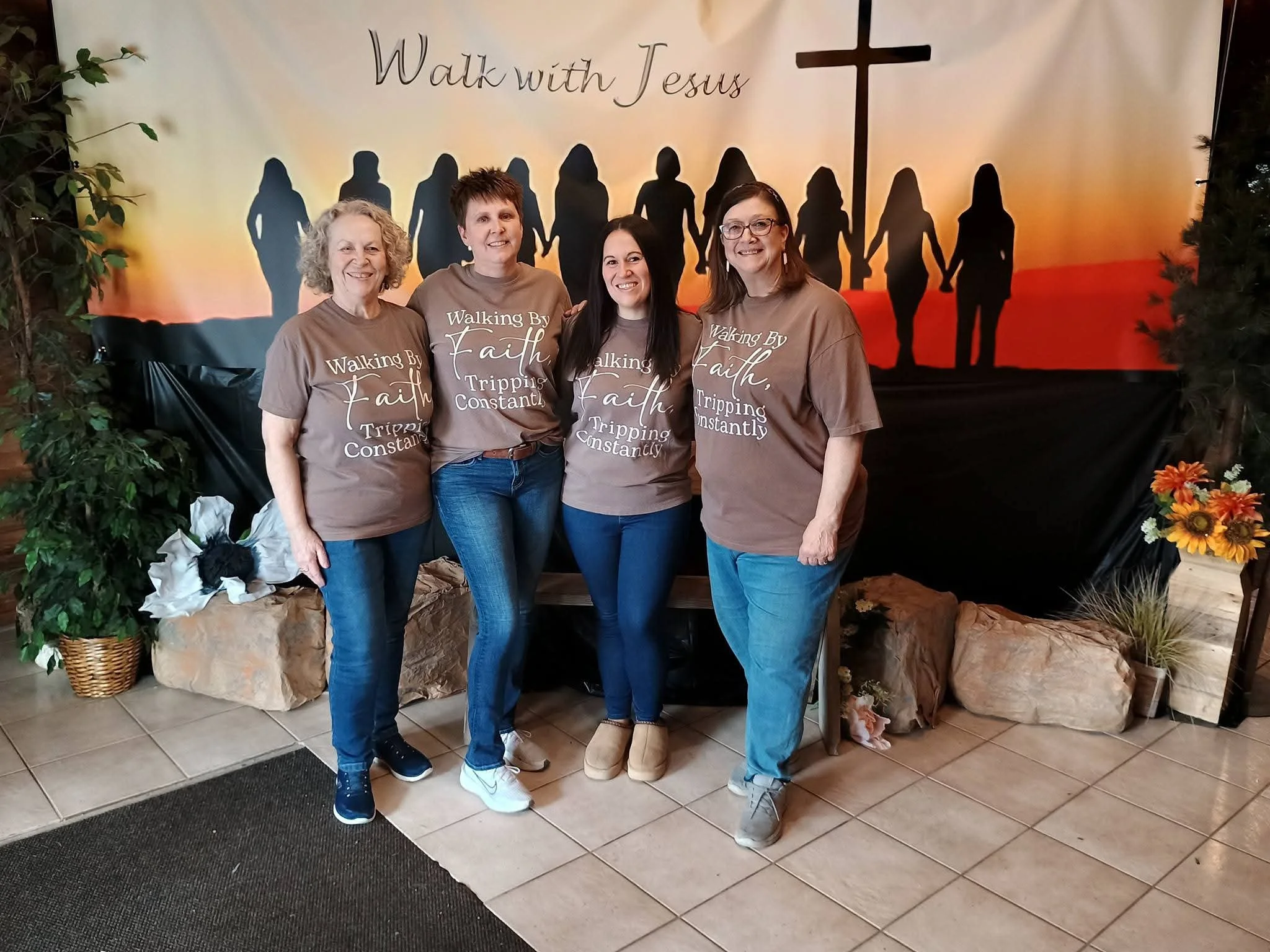 Four women standing inside a church or religious venue with a backdrop featuring silhouettes of people holding hands and a cross. The women are wearing matching brown shirts with the words 'Walking by Faith, Tripping Constantly' printed on them, smil