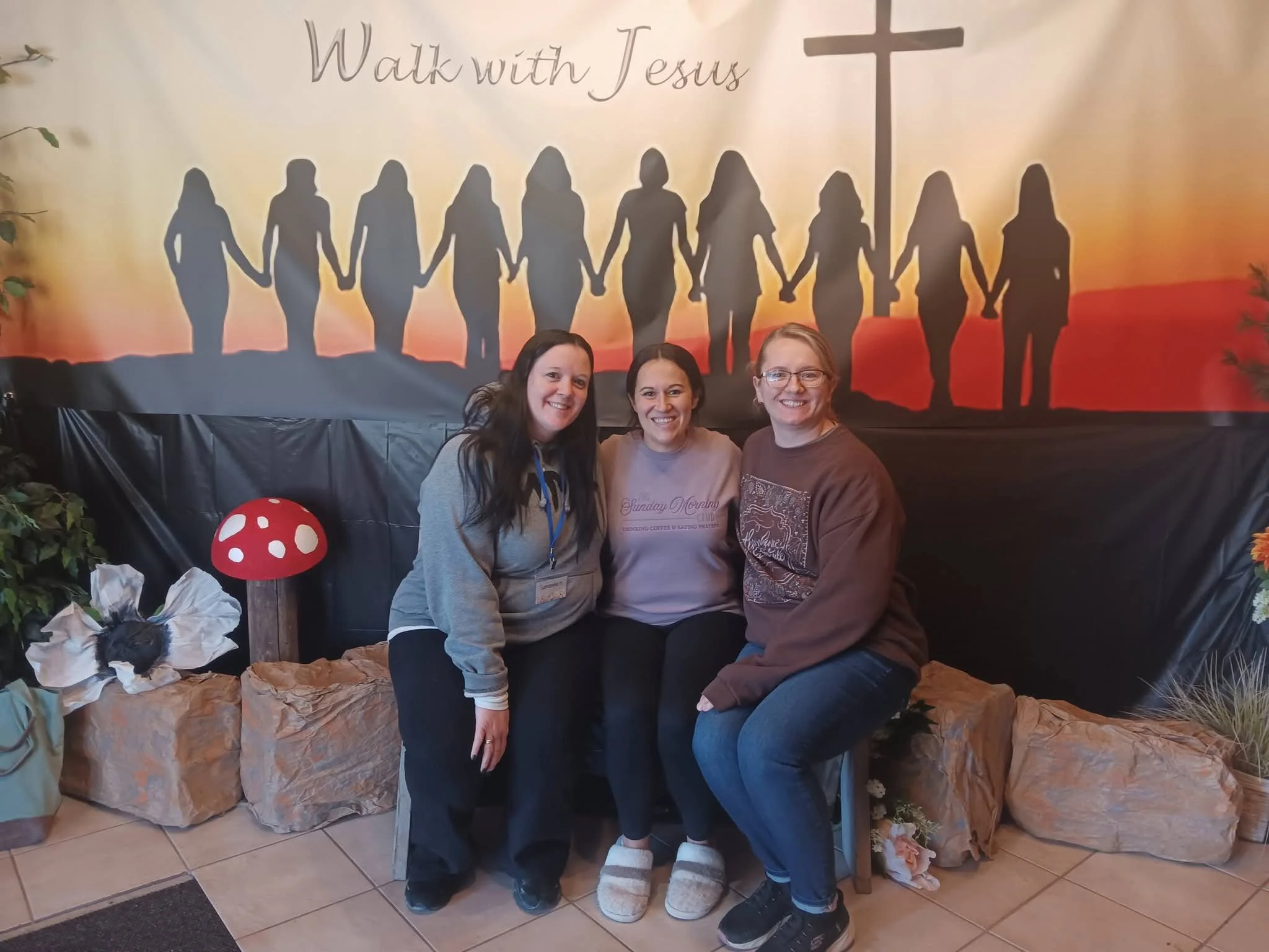 Three women kneeling in front of a large poster that reads "Walk with Jesus" with silhouettes of people holding hands and a cross in the background. There are plants and paper decorations around them.